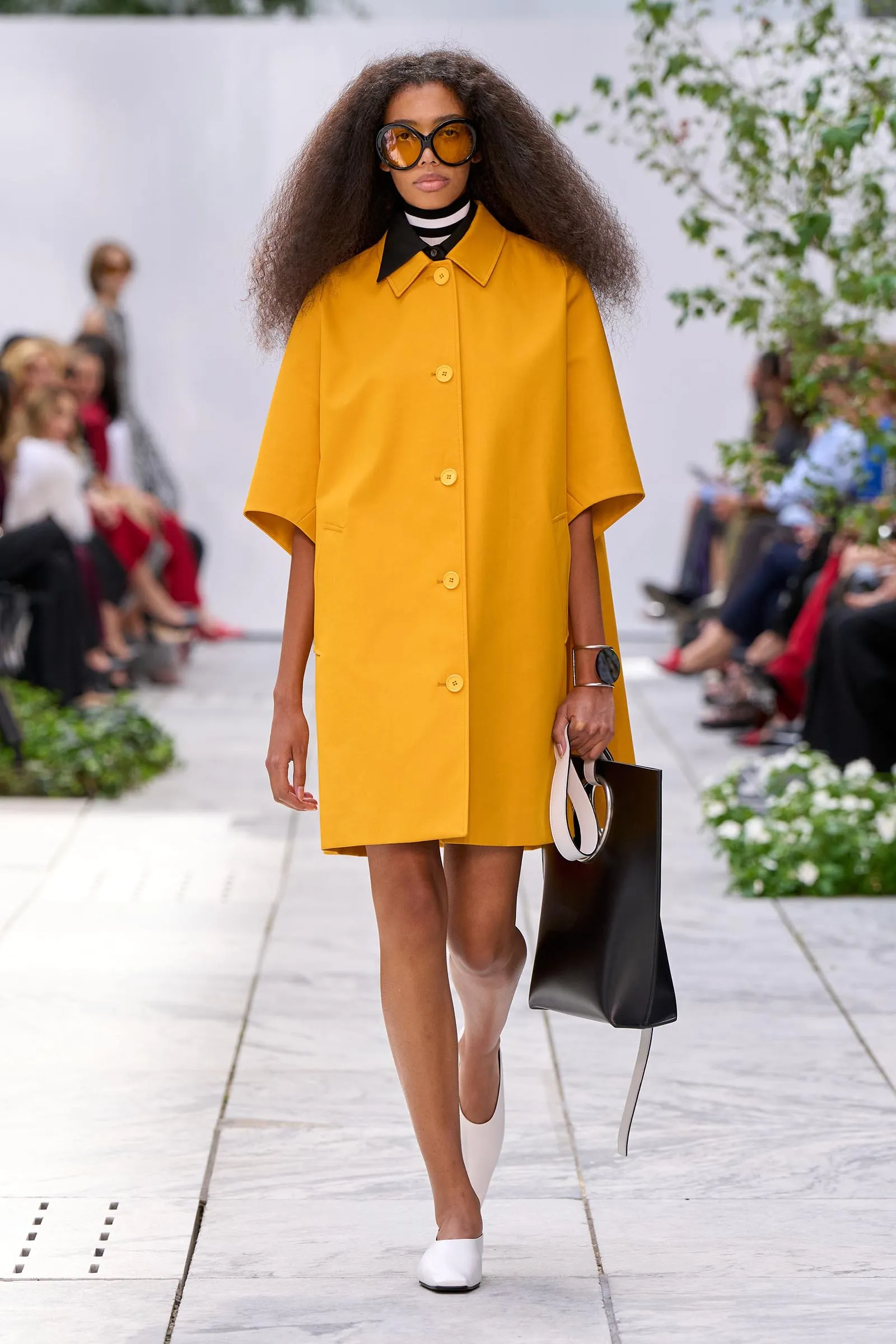 Model wearing a bright mustard-yellow coat with oversized orange sunglasses on a fashion runway, carrying a black-and-white handbag.
