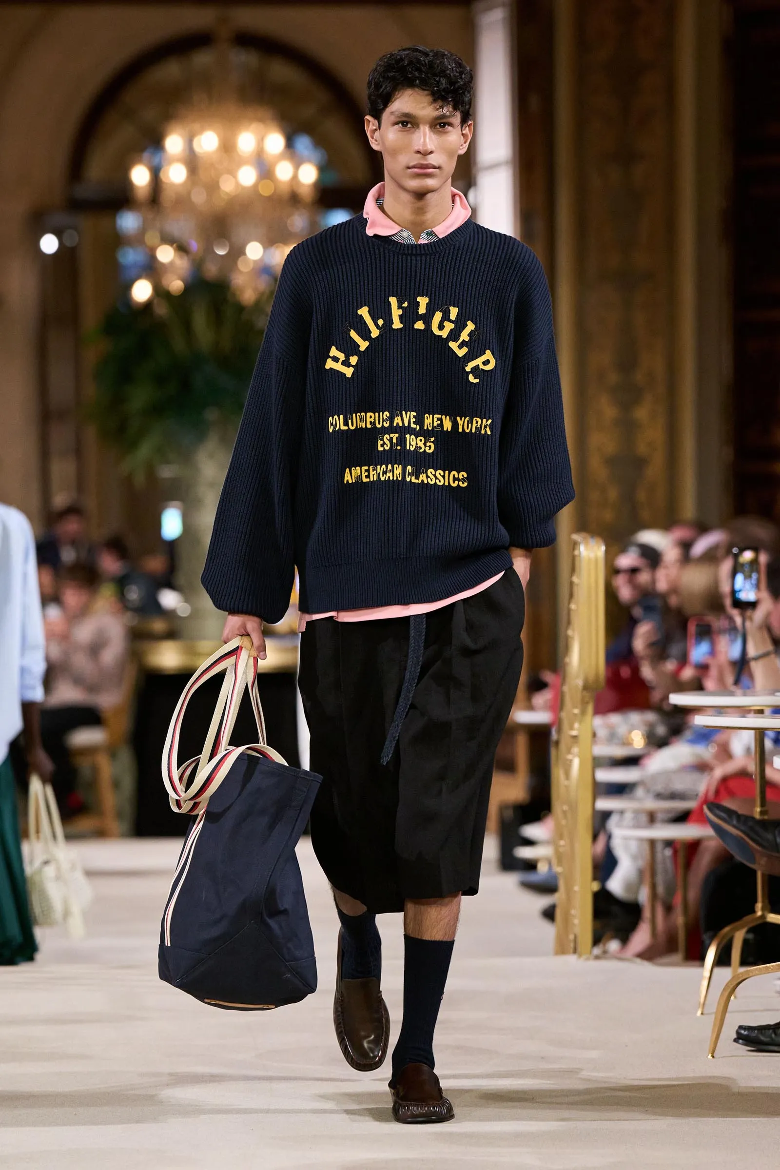 Runway-model wearing a navy knit sweater with yellow lettering, pink collar peeking out, carrying a navy striped tote bag.