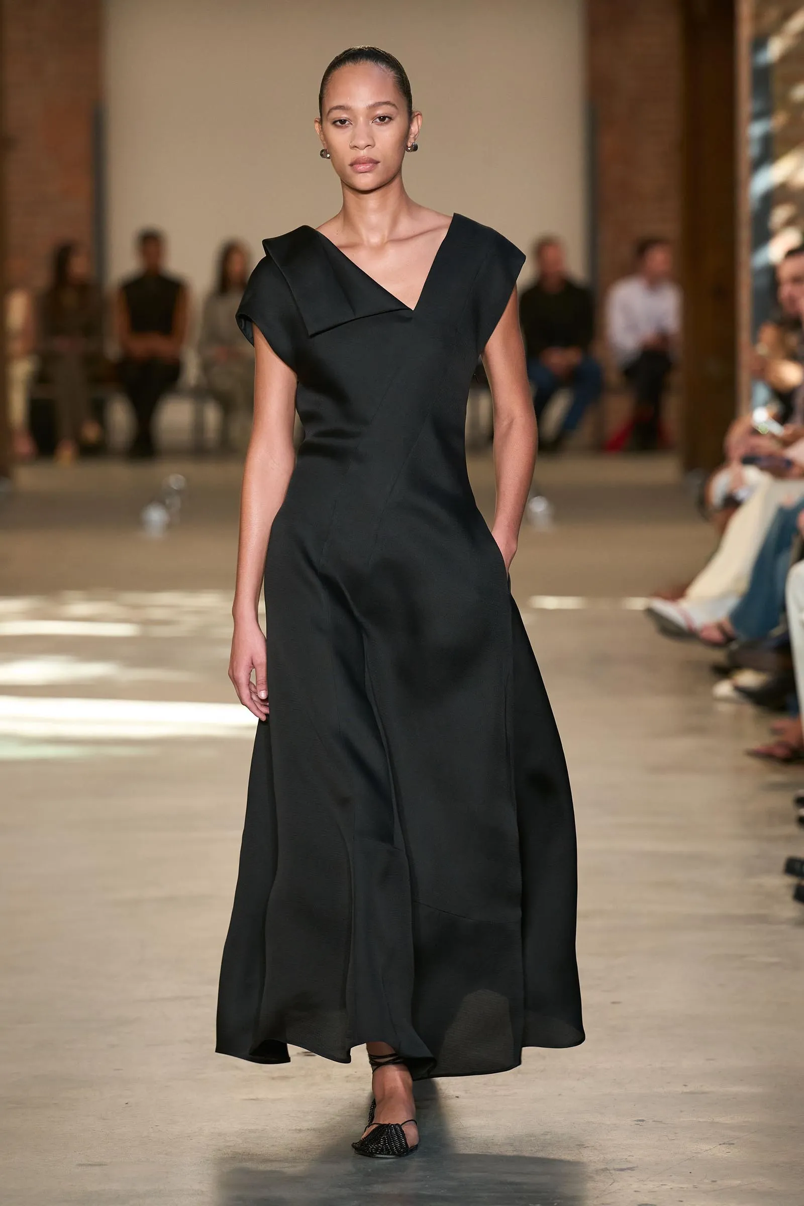 Model walks a fashion runway in a long black dress with an asymmetric neckline; seated audience blurred in the background.