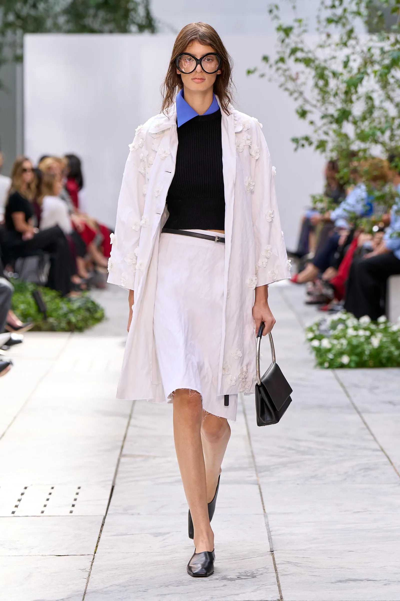Runway model walking in a white floral-embroidered coat over a black top with a blue collar, white skirt, and a black handbag.