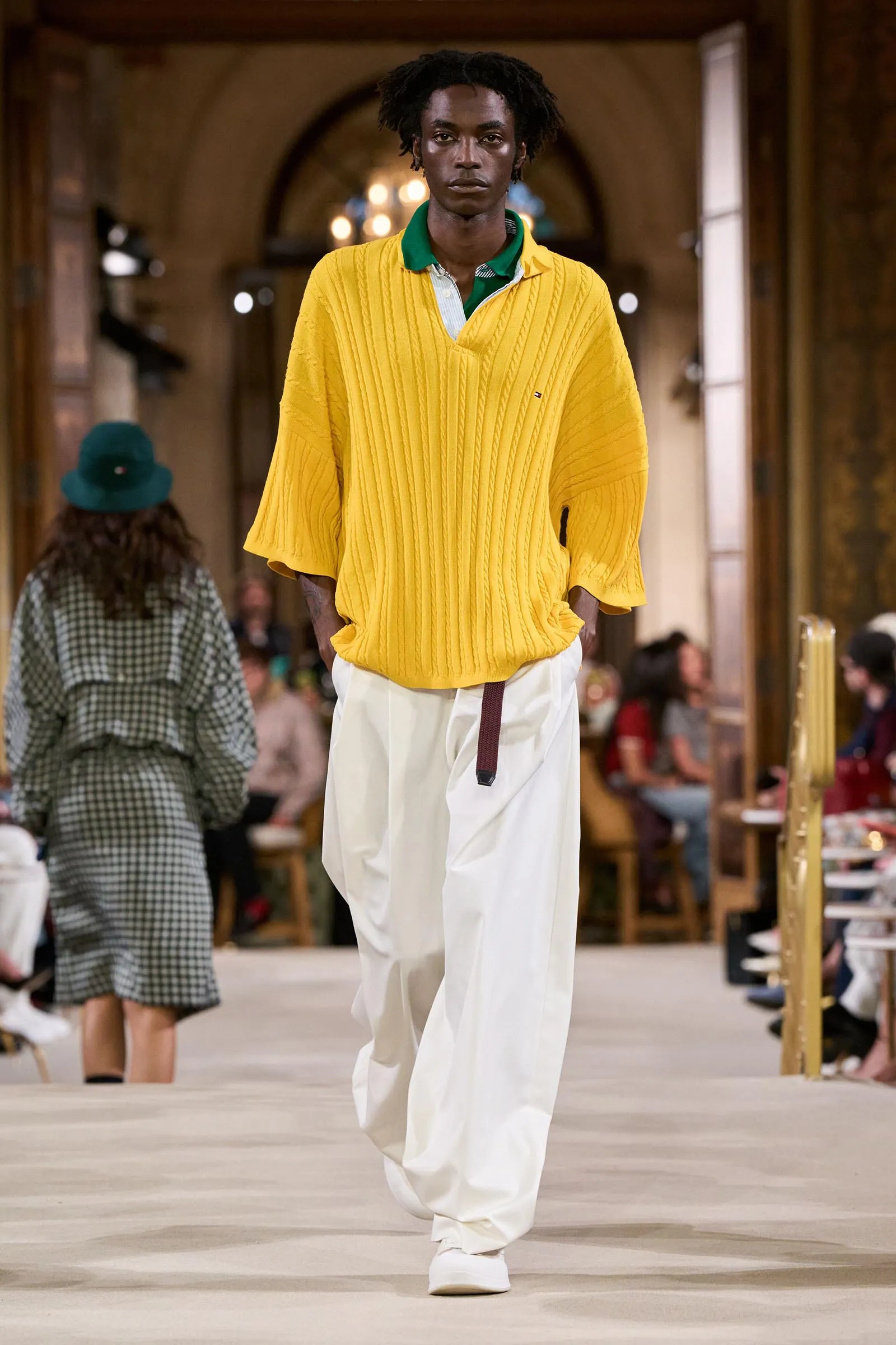 Male model walks a fashion runway wearing a bright yellow oversized cable-knit sweater over a green-collared shirt and white wide-leg pants.