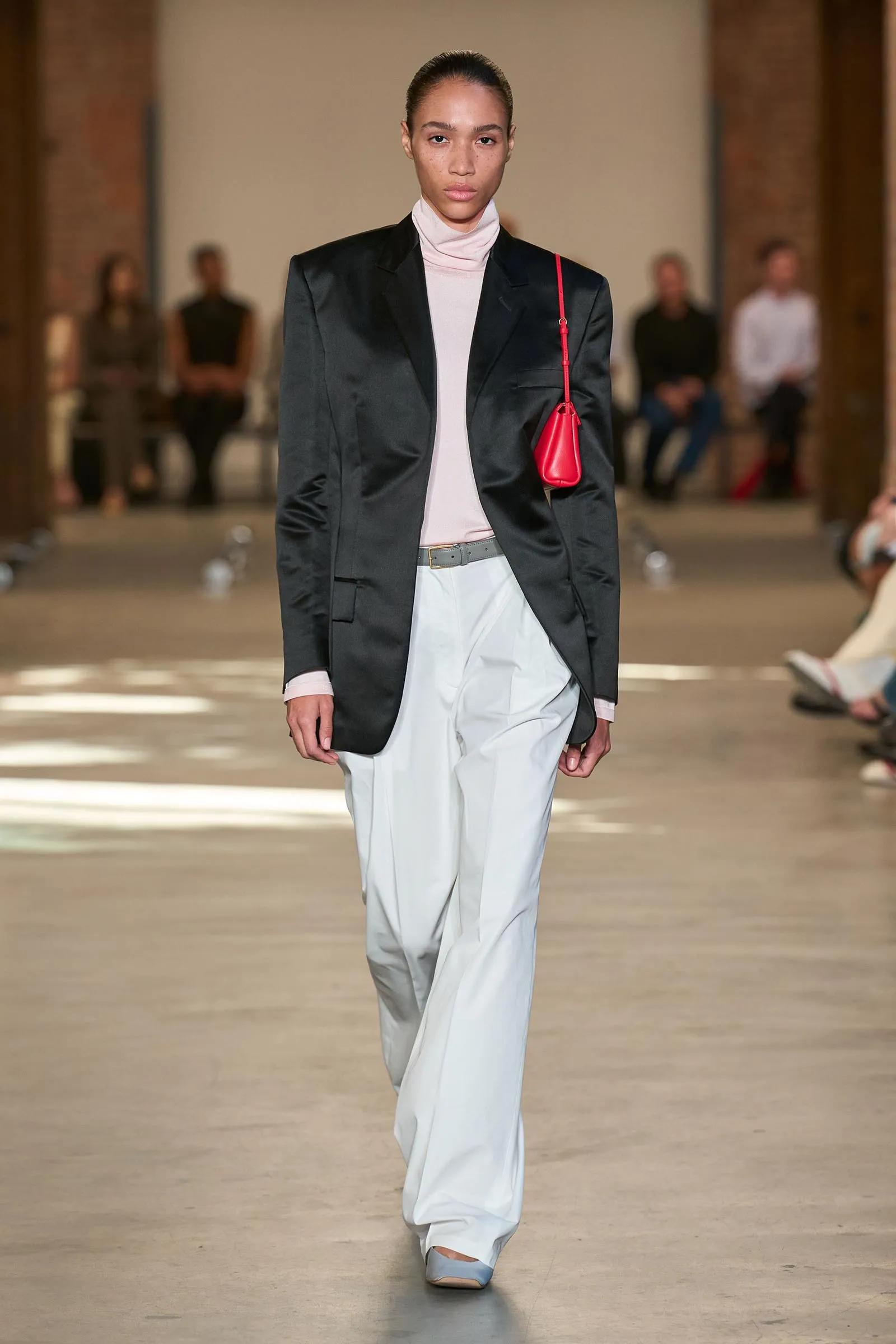 Model walks a fashion show wearing a black blazer, pale pink turtleneck, white wide-legged pants, and a small red shoulder bag.