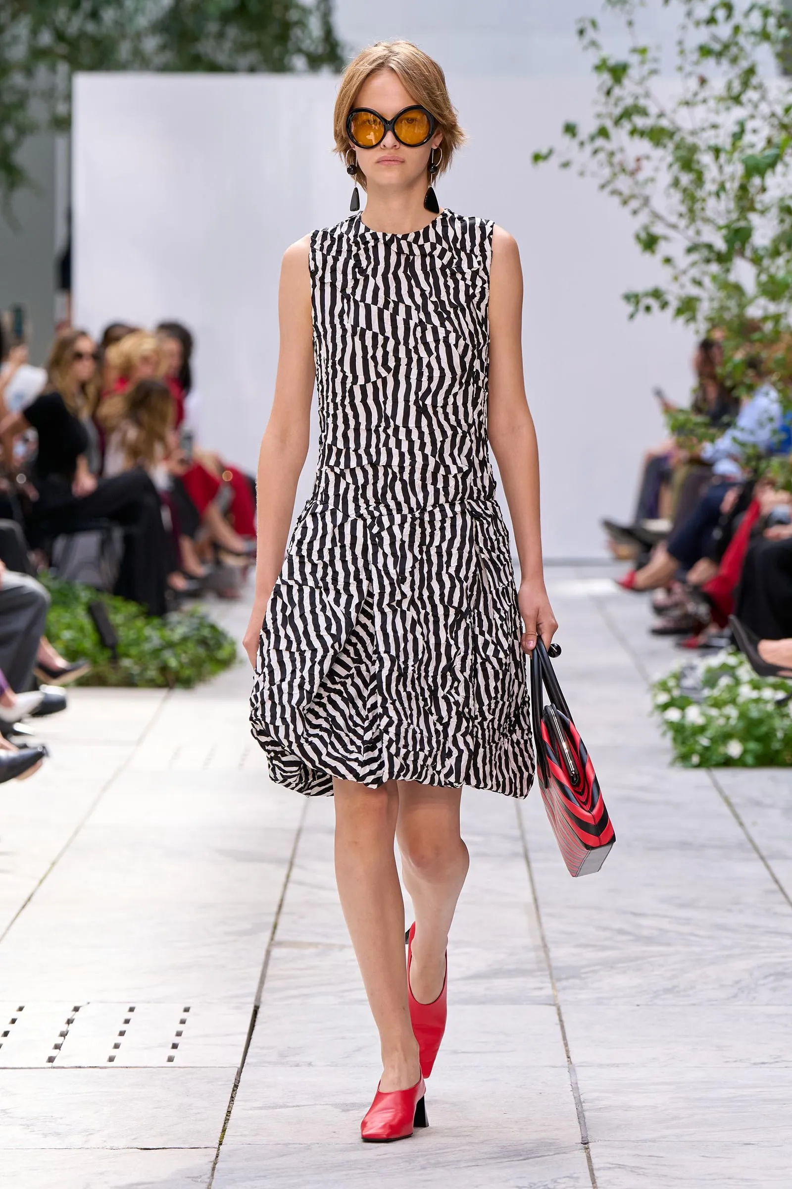 Model walking at a fashion show wearing a black-and-white zebra-print sleeveless dress, orange sunglasses, red heeled shoes, and a red-striped bag.