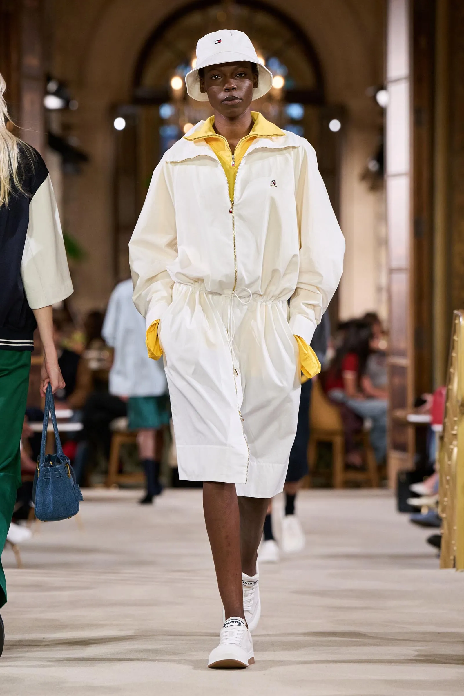Female model walking a fashion runway in a cream windbreaker with yellow accents and a white bucket hat.