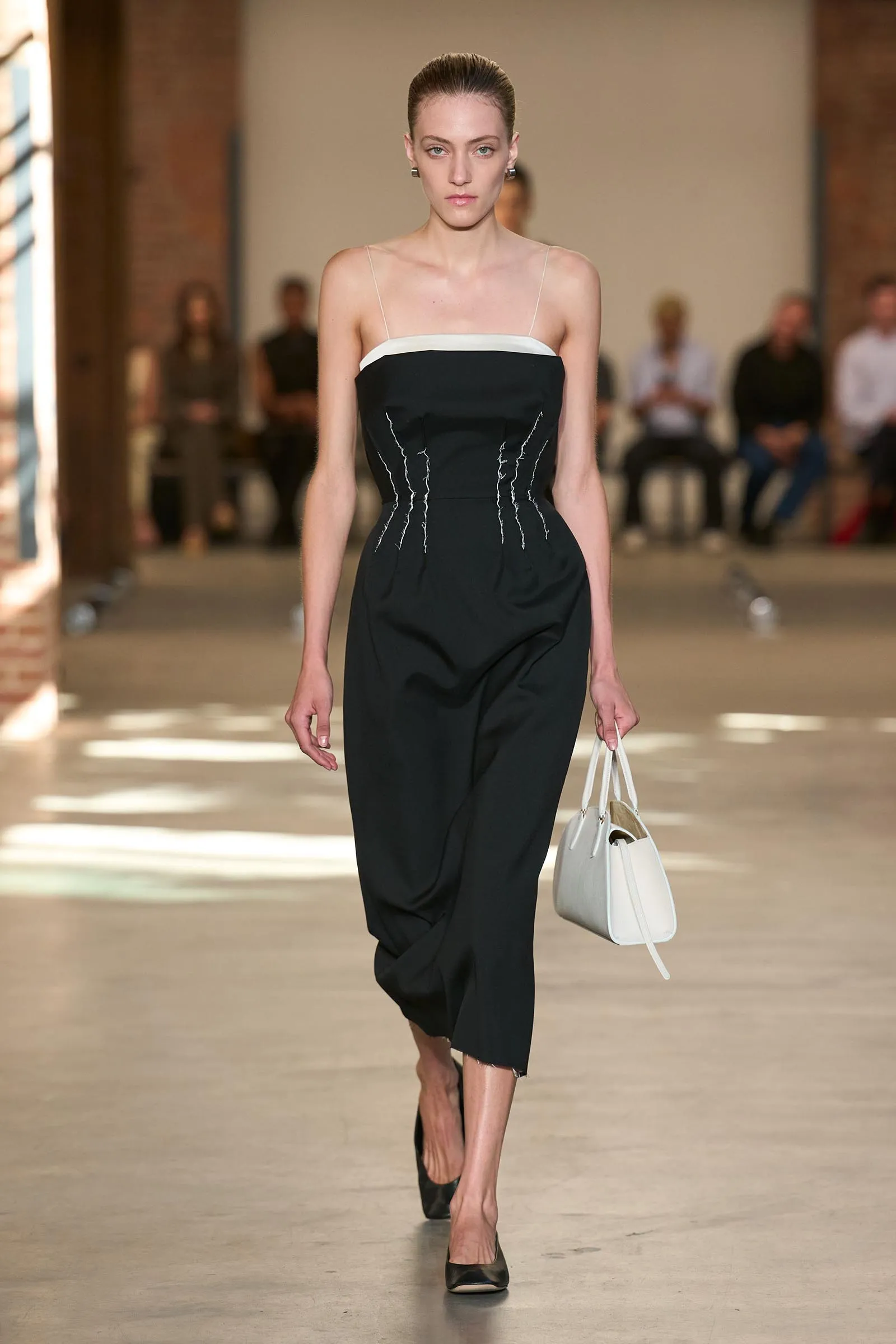 Model walking a fashion runway in a black strapless jumpsuit with white neckline, carrying a white handbag.