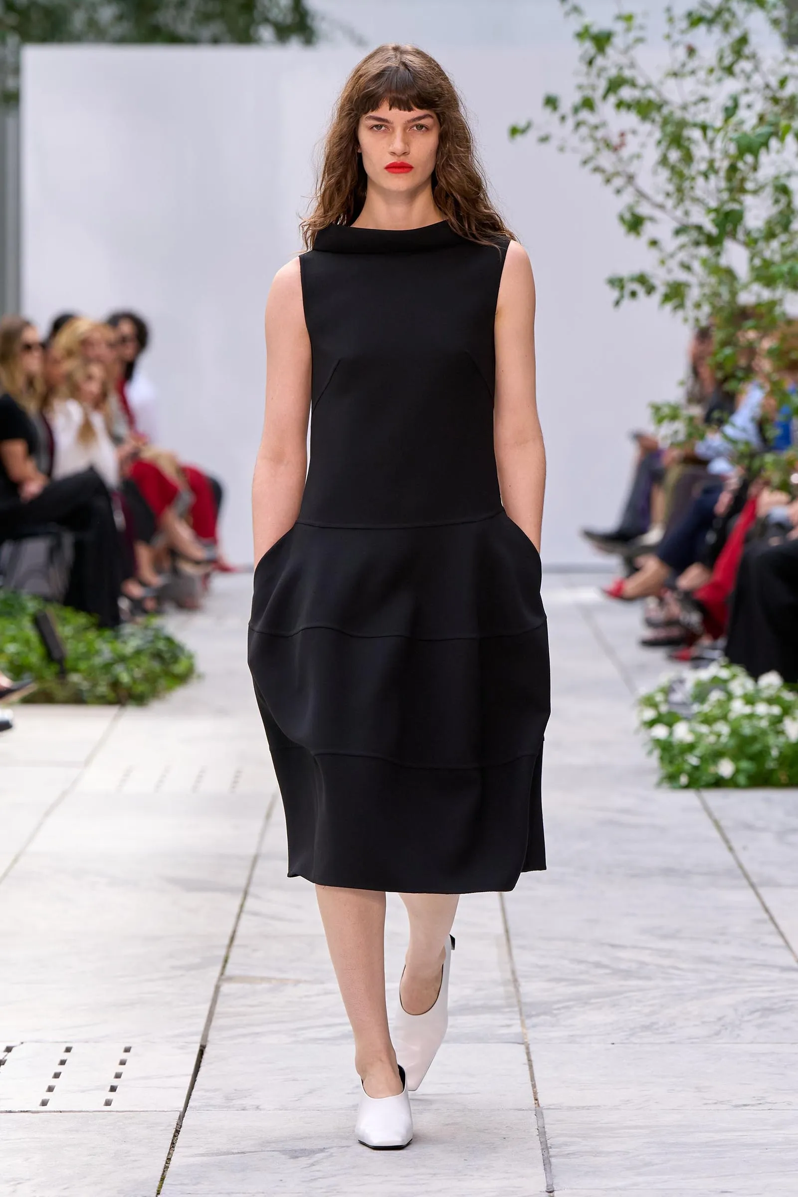 Female model walking a runway in a sleeveless black dress with white flats at an outdoor fashion show, audience seated along the sides.