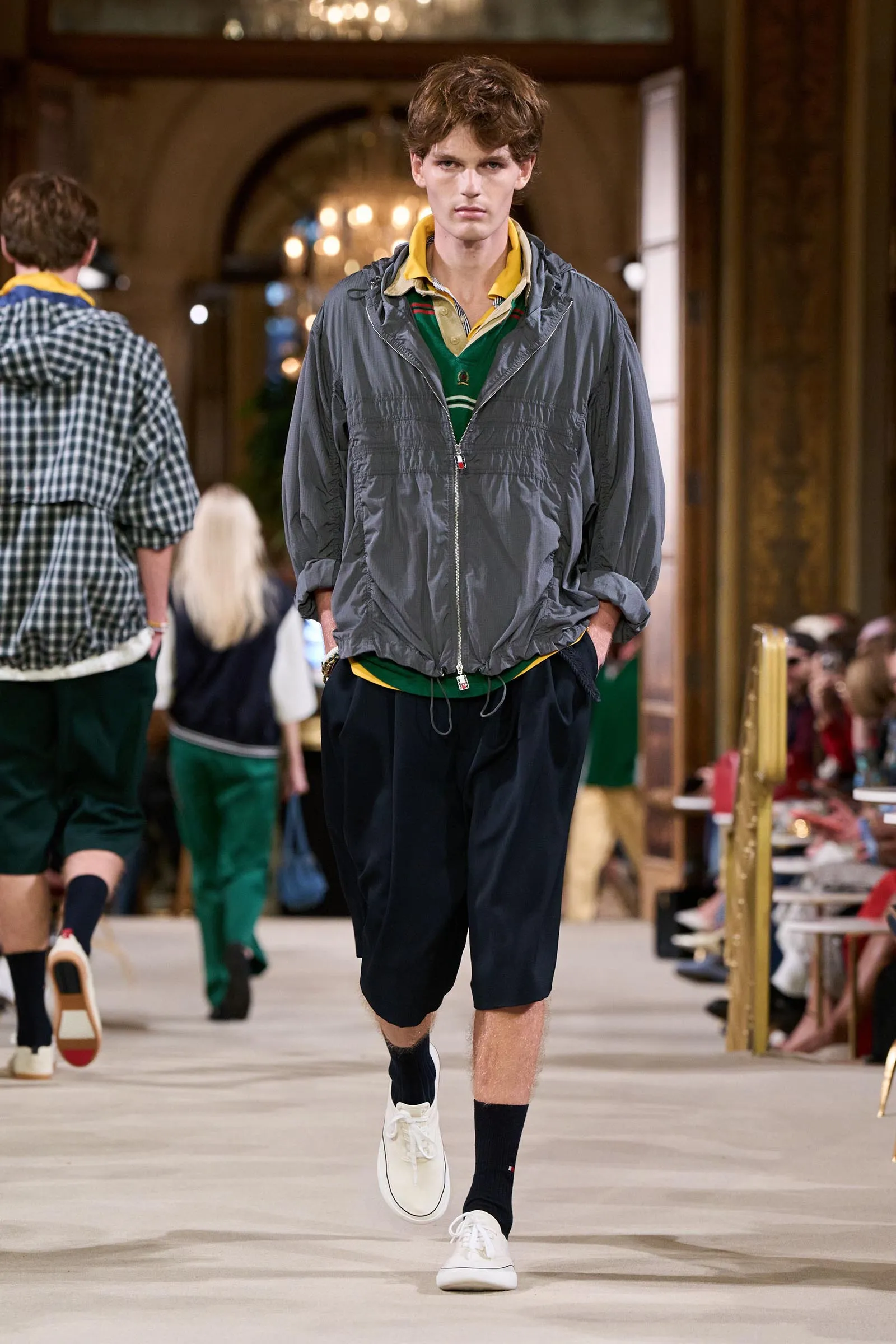 Male model walking a fashion runway in a gray zip-up jacket over a yellow polo and green layers, navy shorts, white sneakers.