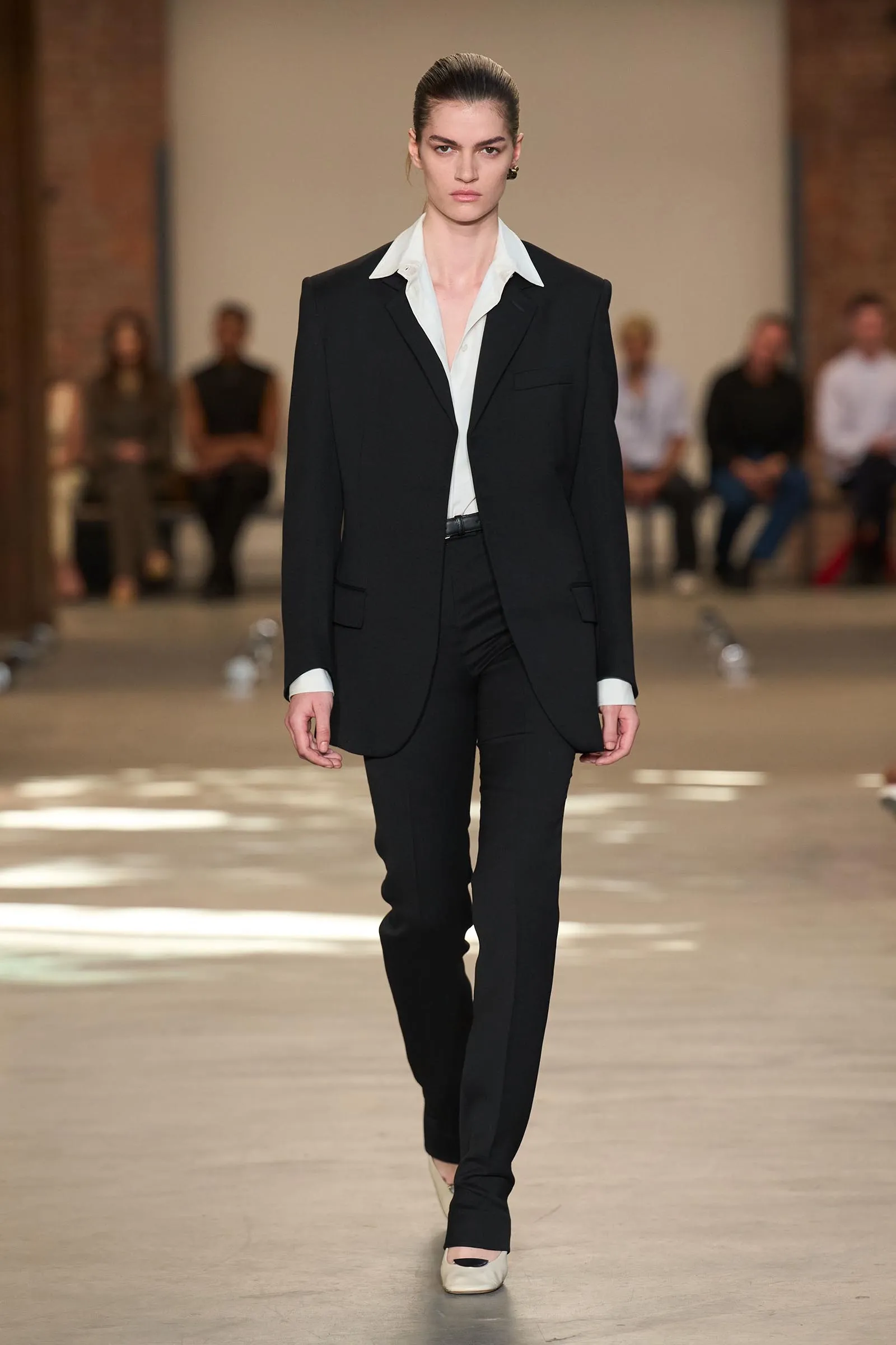 Female model in a black suit with a white collared shirt walking a fashion runway; blurred audience in the background.