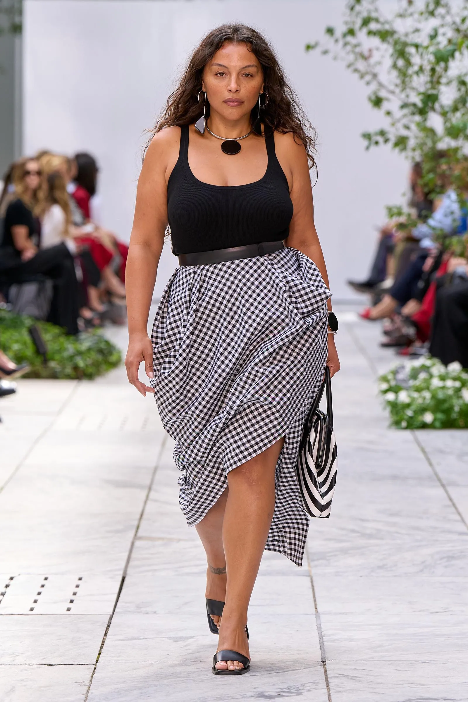 Model walks a runway wearing a black sleeveless top and a black-and-white gingham wrap skirt with a wide belt and accessories.