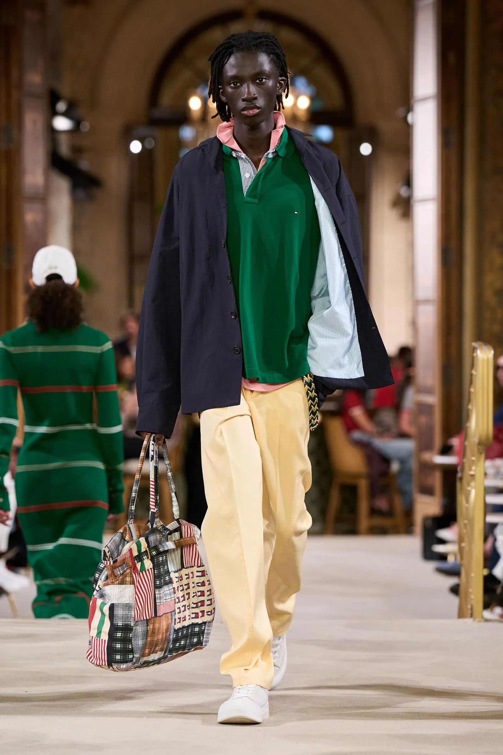 Male model walking a runway in a green polo with a pink-collar, navy coat, beige pants, white sneakers, and a large patchwork tote bag in hand.