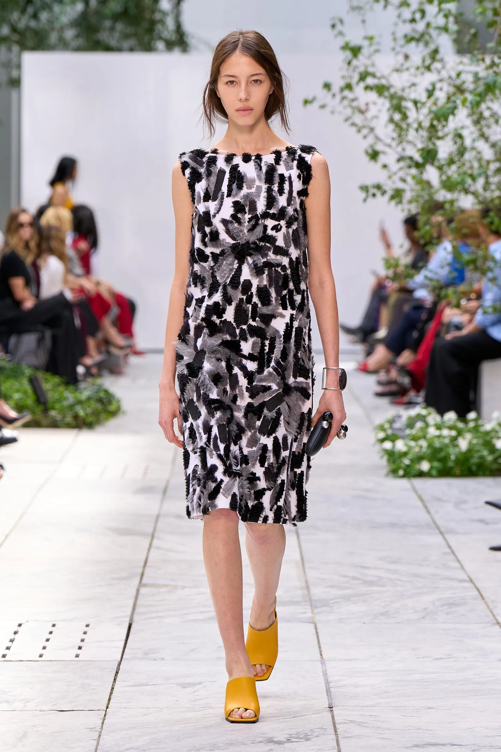 Model walks a runway in a black-and-white feather-patterned sleeveless dress with yellow sandals, audience blurred in the background.