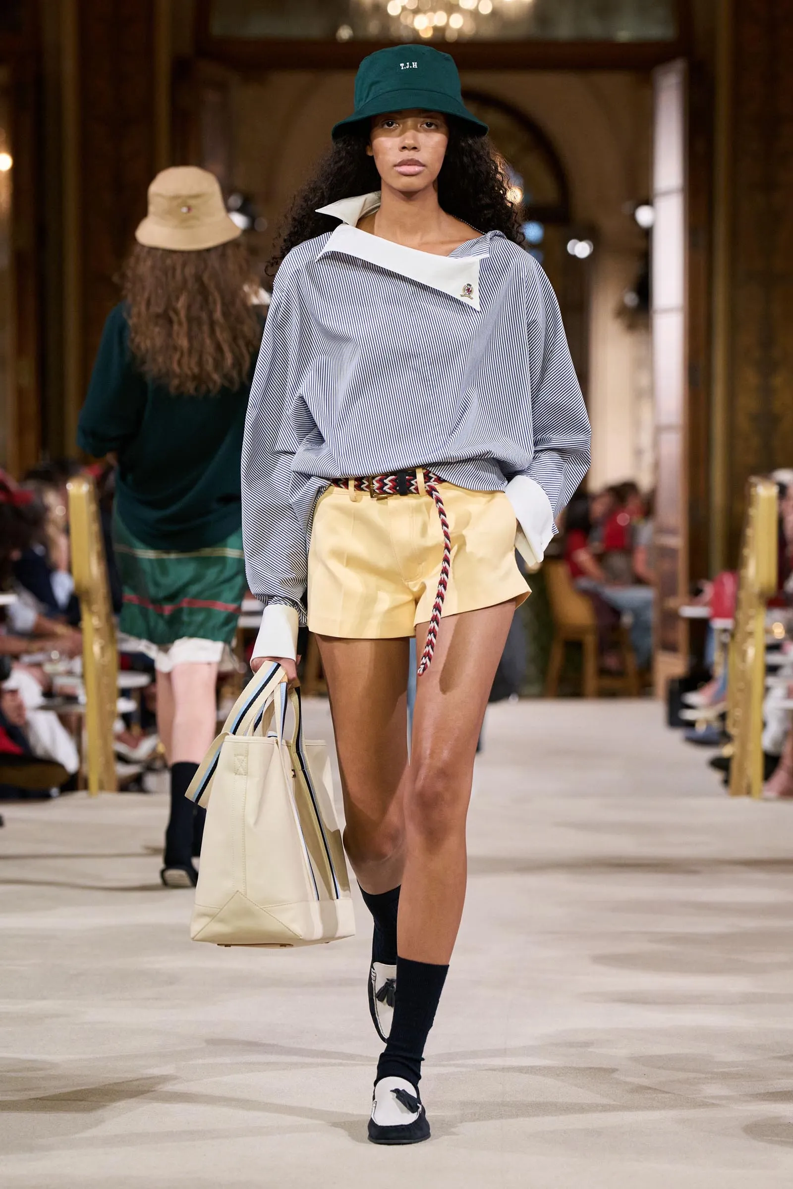 Runway model walking in a blue striped oversized blouse, pale yellow shorts, green bucket hat, and a beige striped tote bag.