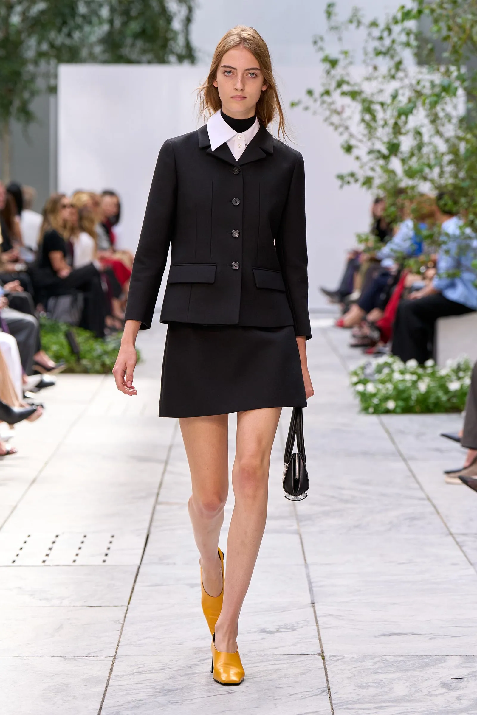 Runway model wearing a black tailored blazer and skirt with a white collared shirt, yellow ankle boots, and a small black handbag, walking down the runway.