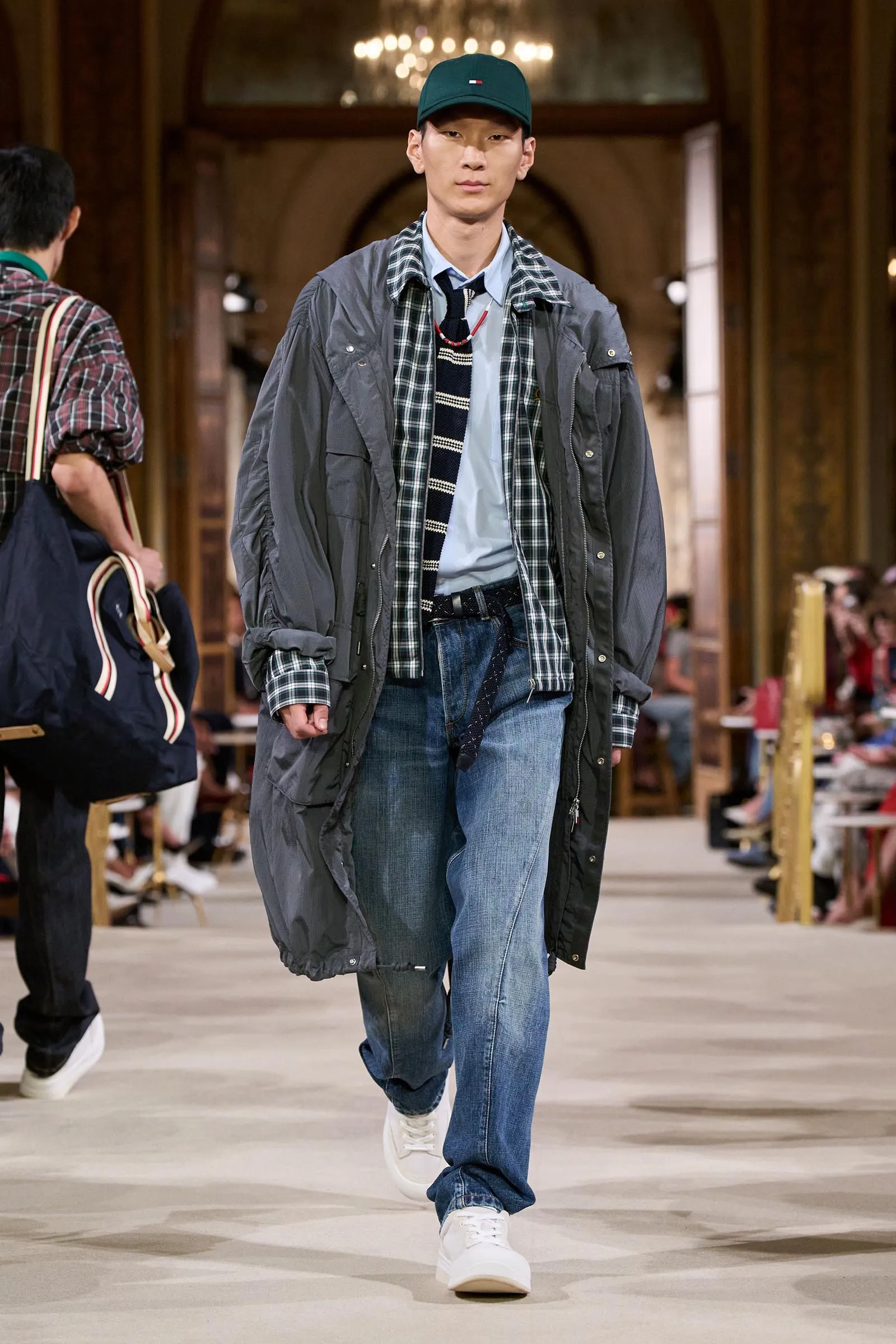 Male model walking a fashion runway in an oversized gray coat over a plaid shirt, light blue tee, striped tie, and blue jeans, wearing a green cap and white sneakers in a grand hall setting.