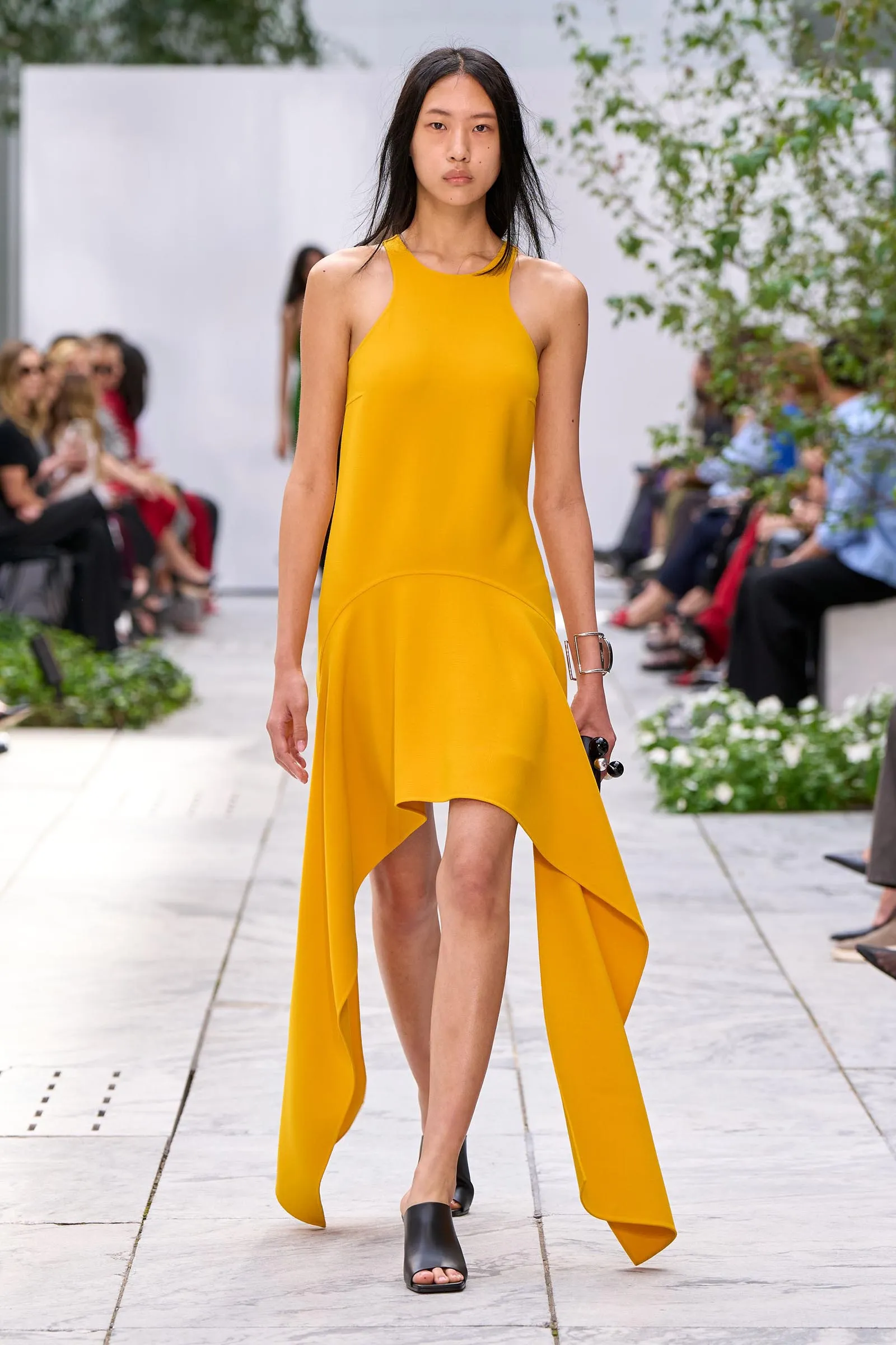 Model walking a fashion runway in a bright yellow high-low dress with spectators along the sides in a blurred background