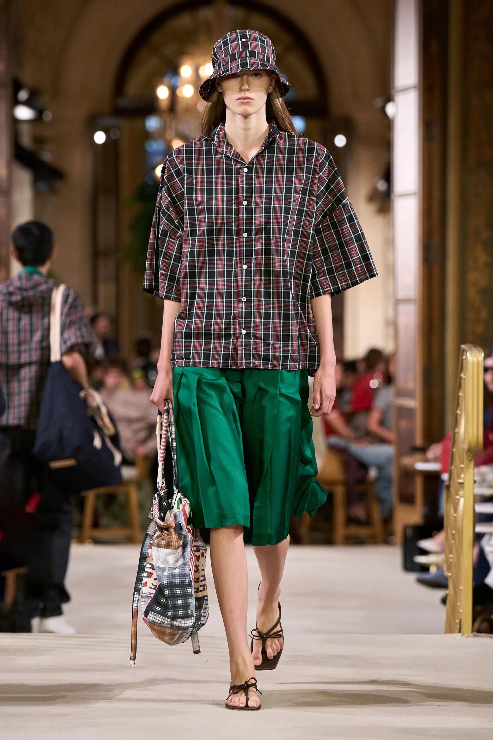 Model walks a fashion runway wearing a black-and-red plaid oversized shirt and matching bucket hat, green wide shorts, and sandals while carrying a patterned bag.
