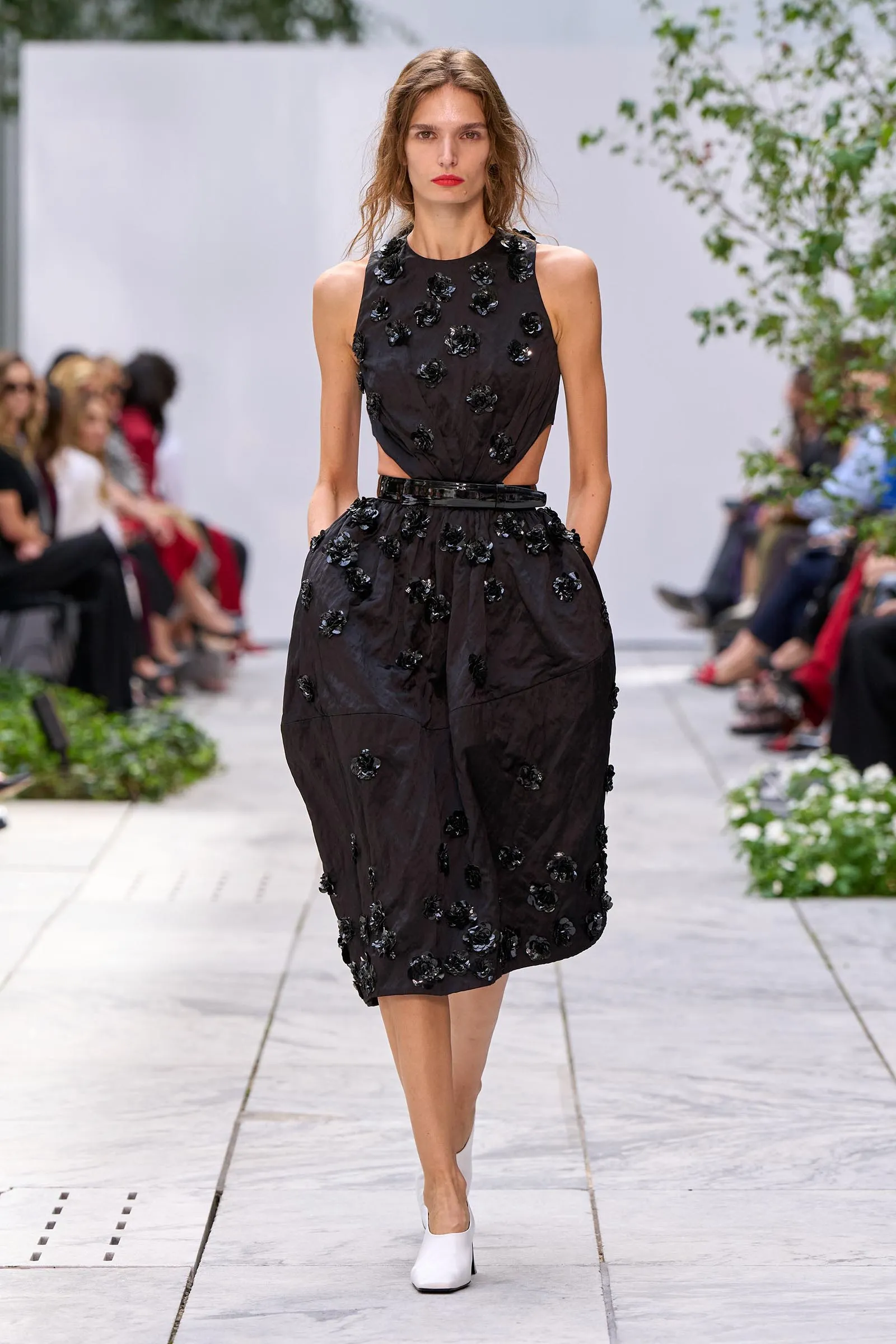 Model walks the runway in a sleeveless black dress with scattered black floral appliques, belted at the waist, paired with white ankle boots.
