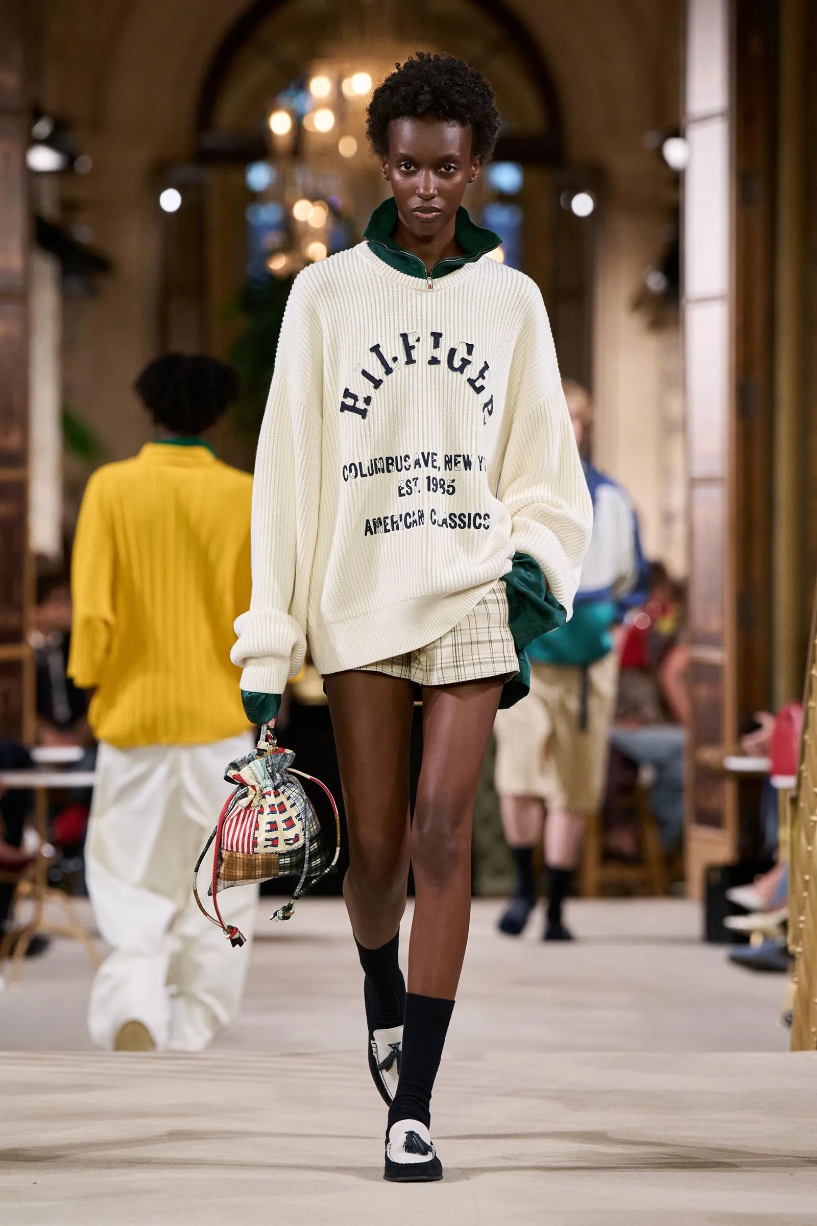 Runway model walking toward the camera in a cream oversized knit sweater with a green collar, plaid shorts, and a patterned handbag, in a busy indoor venue.