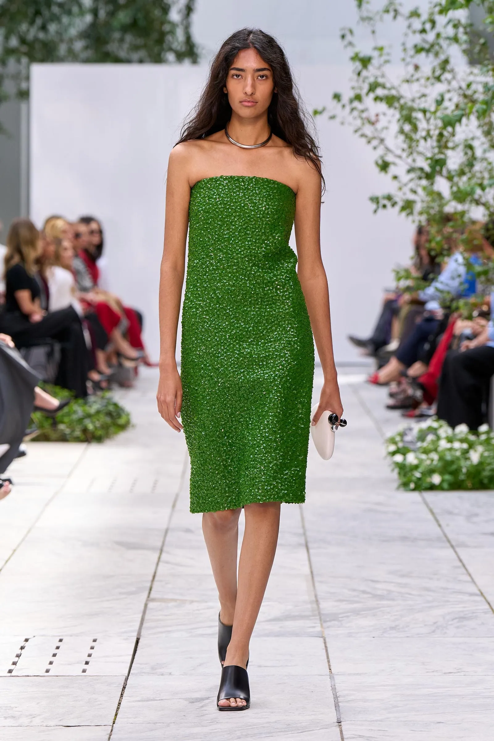 Model walking a runway in a strapless emerald green textured midi dress, with a silver necklace and white clutch in hand.