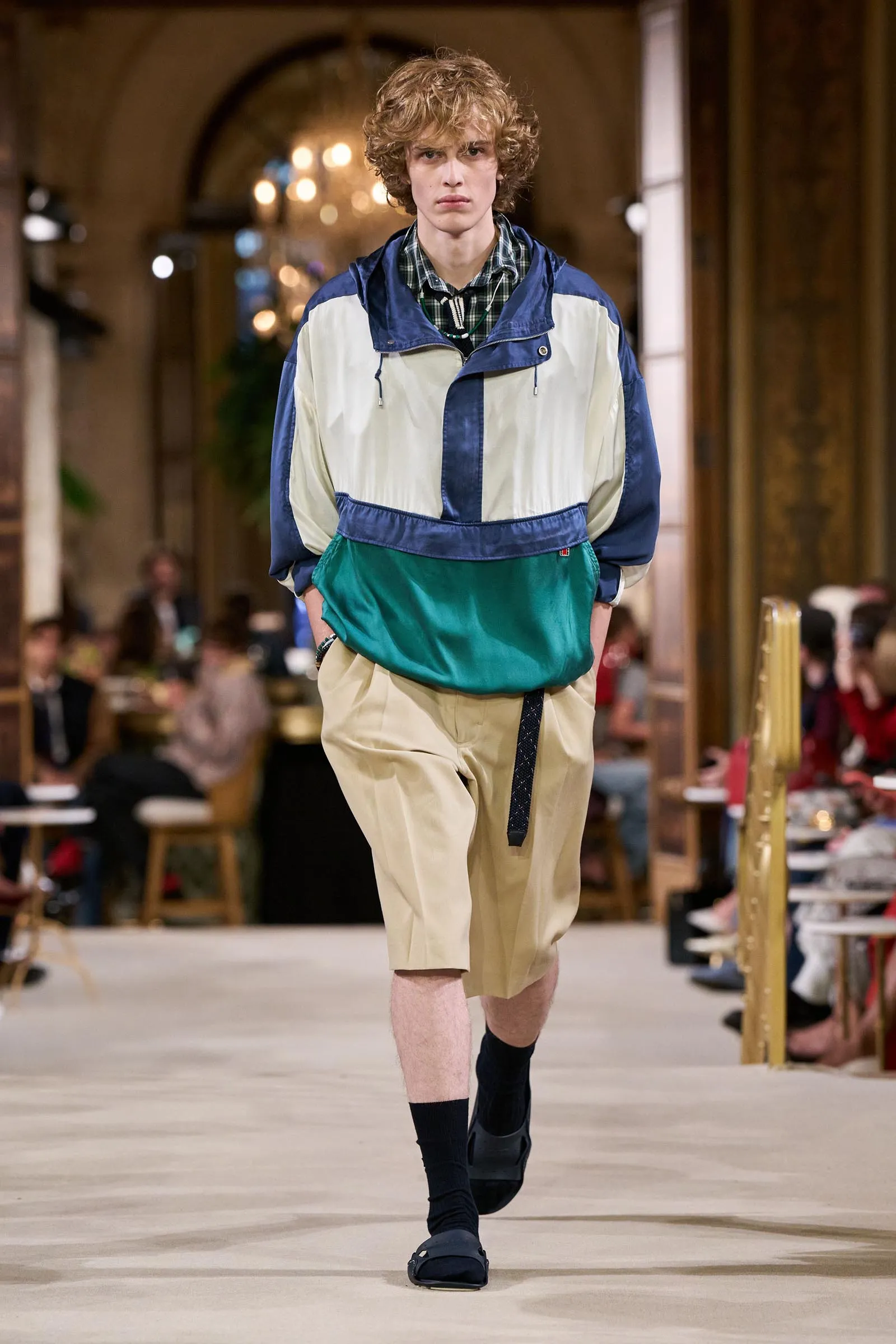 Male model walks the runway in a color-block windbreaker over a plaid shirt, beige shorts, and navy socks.