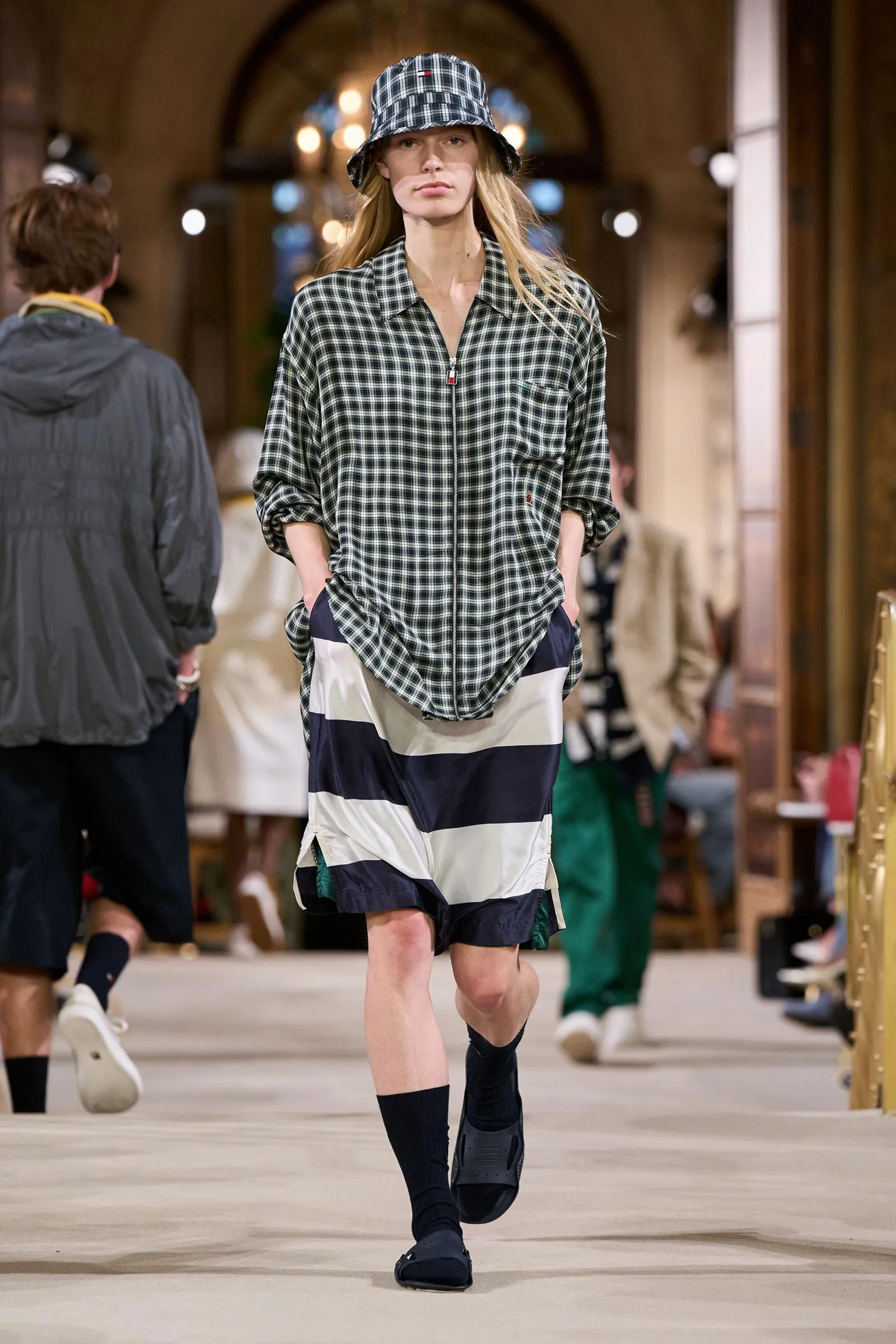 Model walks a fashion show wearing a black-and-white plaid zip-front shirt and bucket hat, with navy-white striped shorts, hands in pockets in a warm-lit venue.