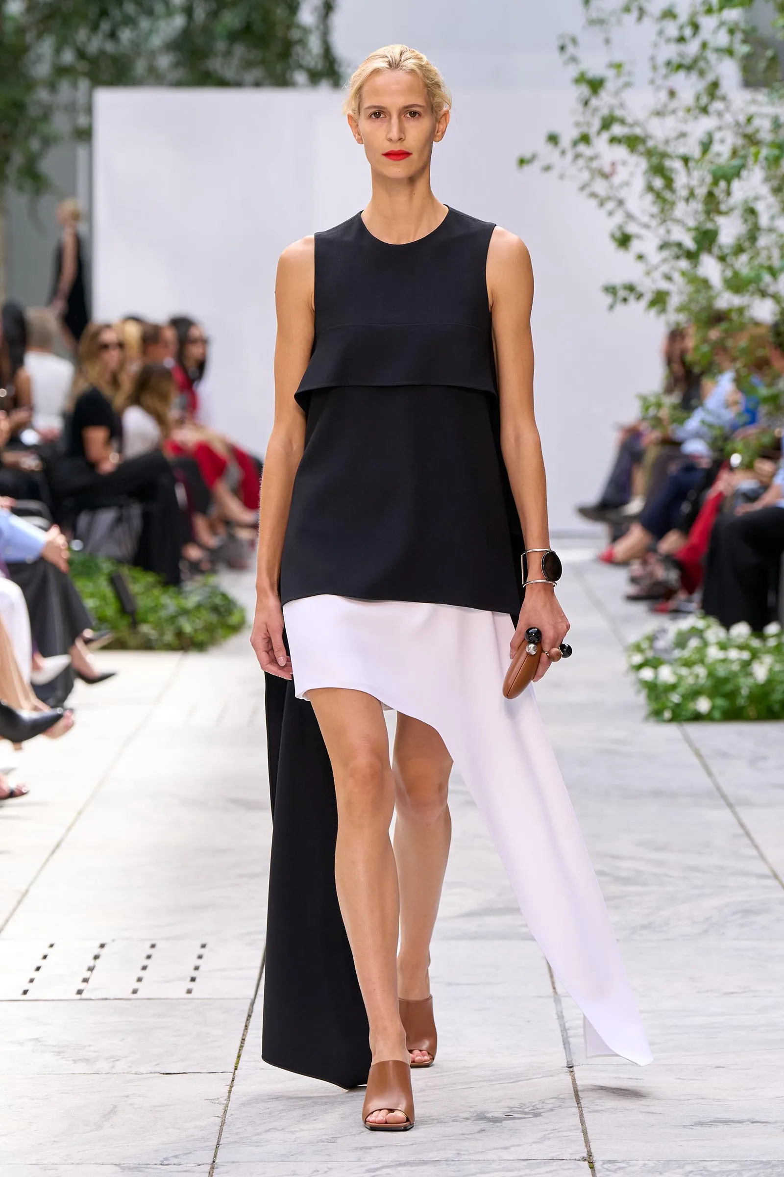 Full-length runway image of a model wearing a sleeveless black top with a layered, color-blocked black-and-white bottom and brown sandals, walking the catwalk with audience in the background.