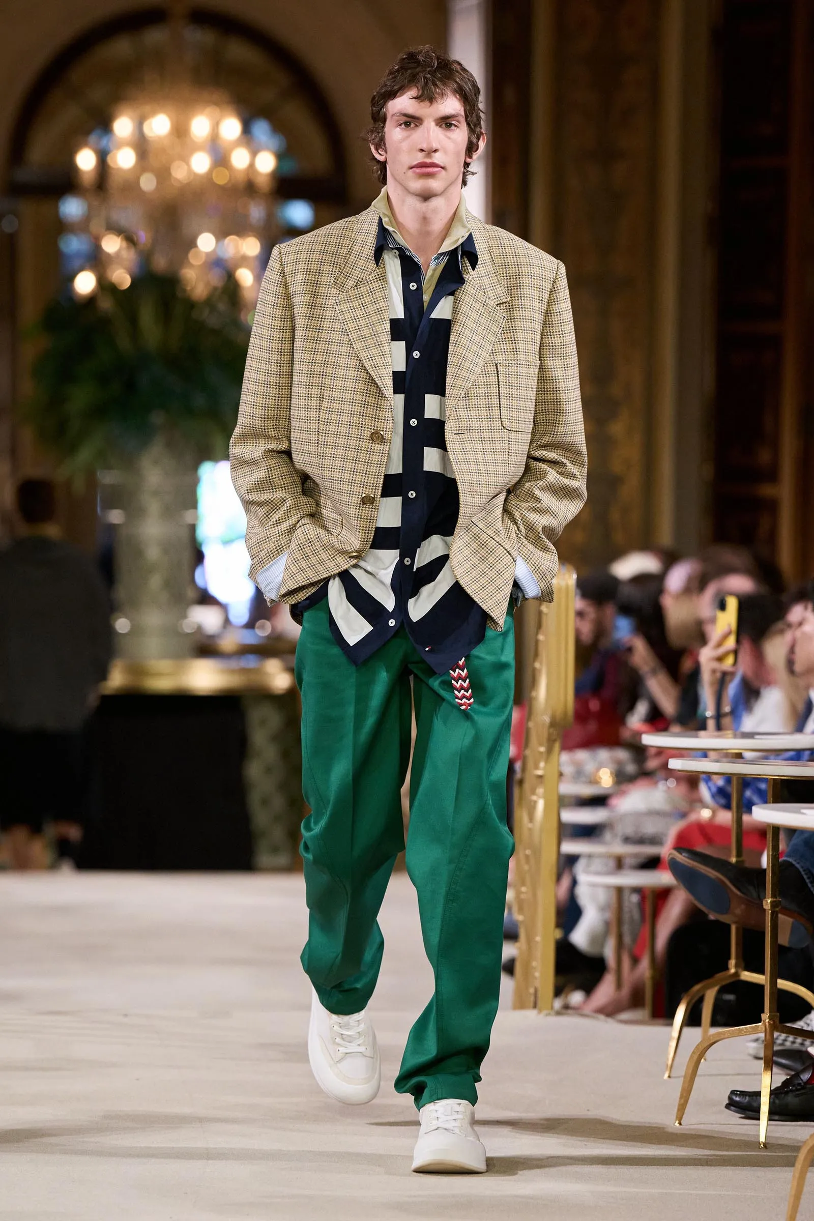 Male model walking a runway in a beige check blazer, black-and-white striped shirt, bright green pants, and white sneakers.