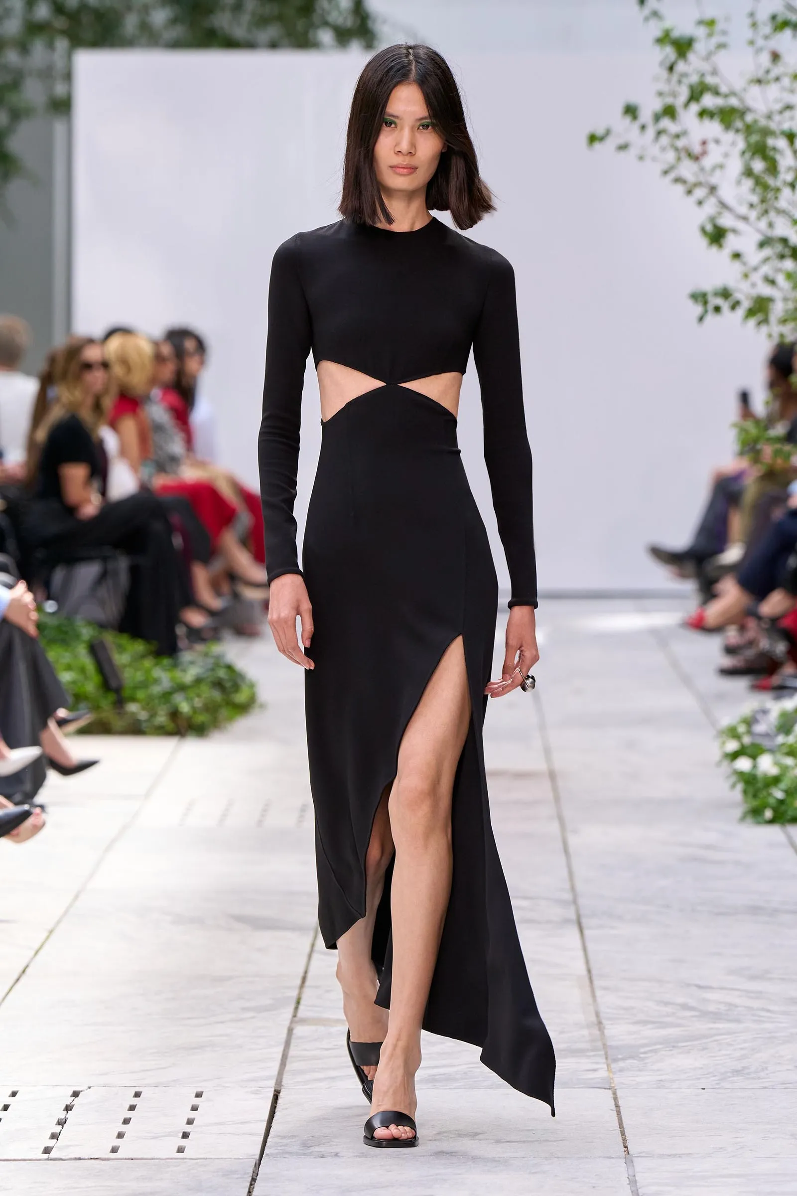 Runway model in a black long-sleeve gown with cutouts at the waist and a high slit, walking a fashion show runway.