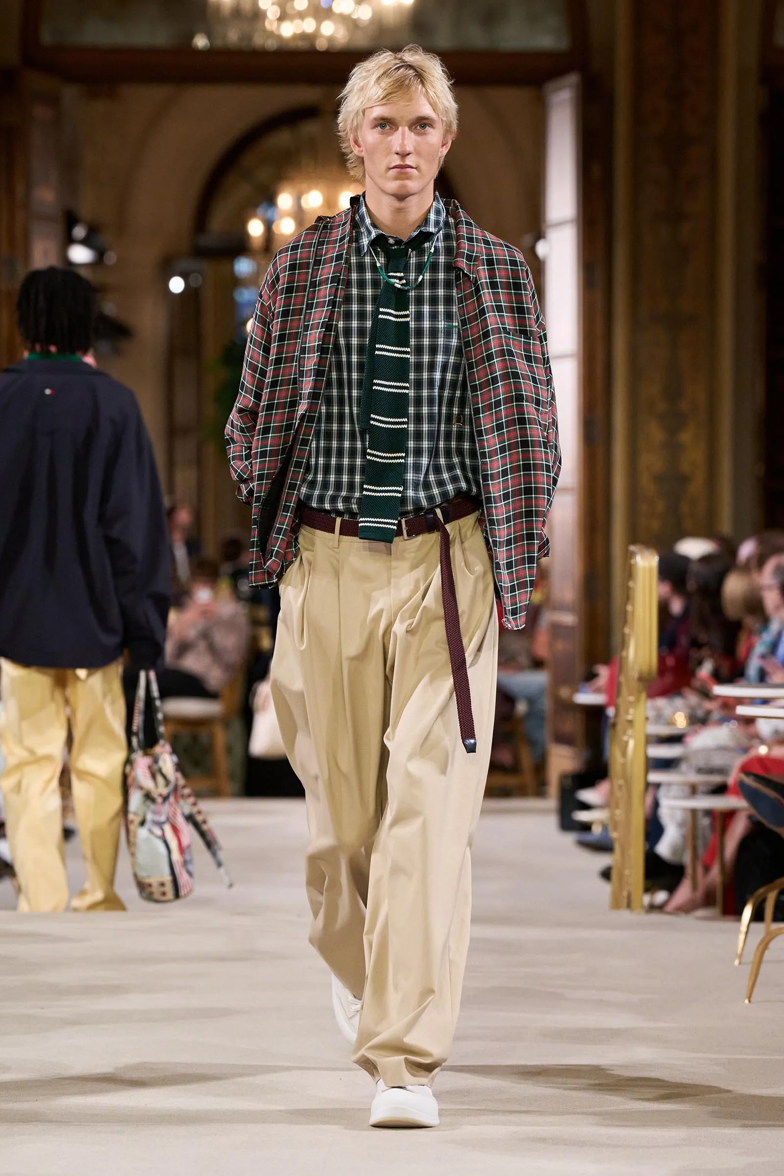 Male model walks a fashion runway wearing a red plaid jacket over a green plaid shirt with a green striped tie and beige baggy trousers, white sneakers.