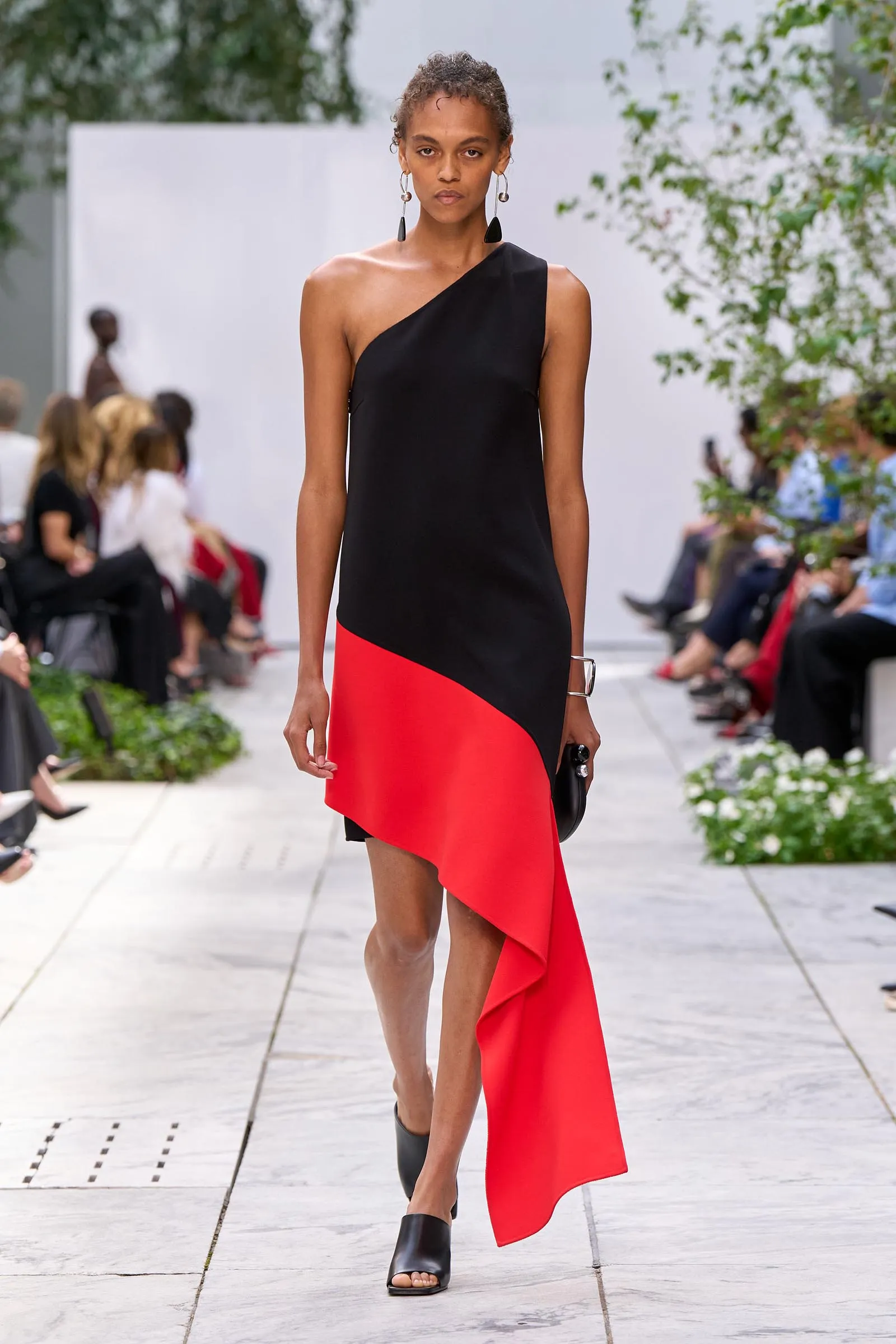 Model walking a runway in a black one-shoulder dress with a red diagonal panel, paired with bold earrings and black sandals on an outdoor catwalk.