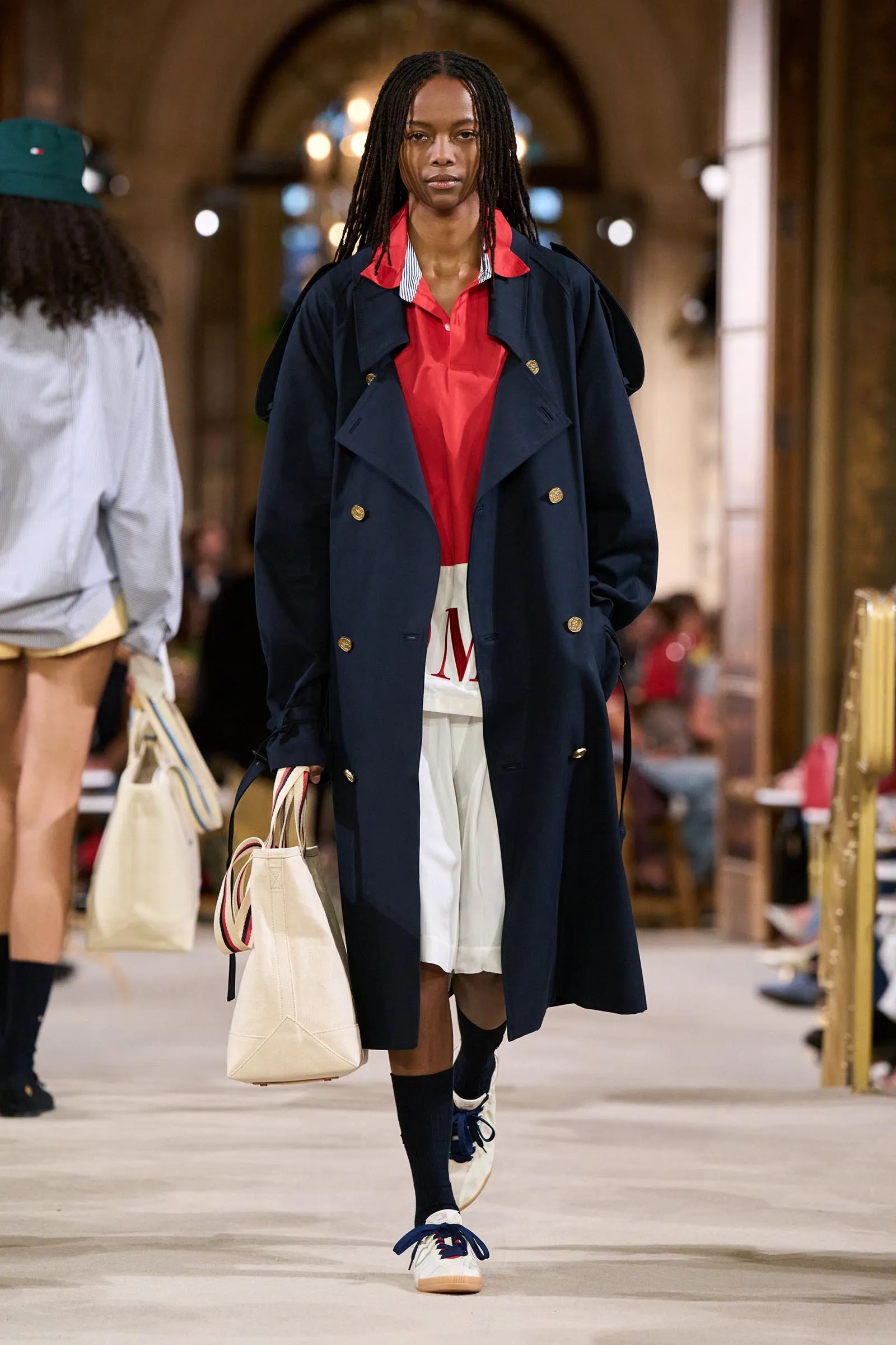 Model walking a fashion runway in a navy trench coat over a red shirt, white shorts, black socks, and white sneakers with blue laces, holding a beige tote bag.
