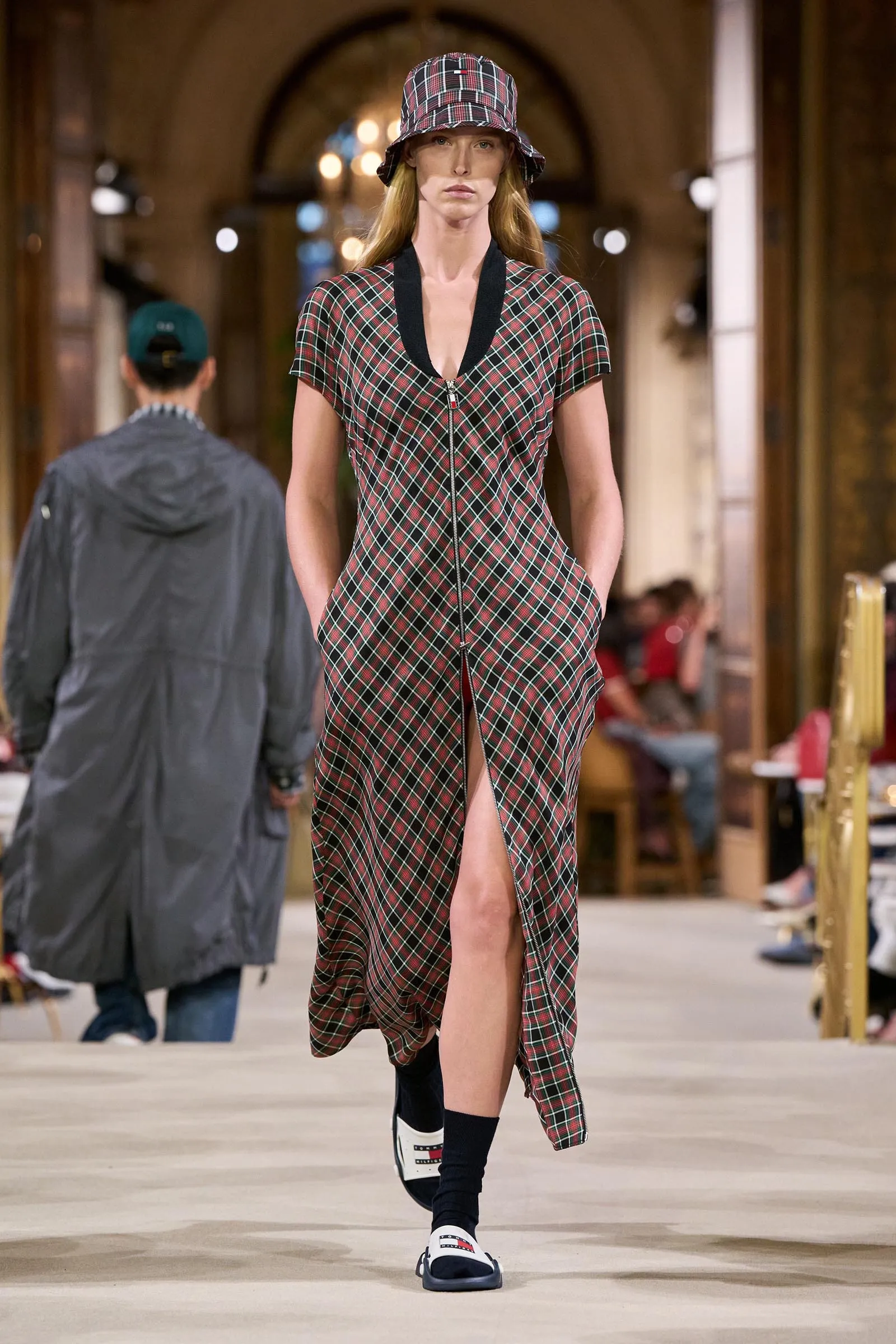 Model walks the runway in a red-green plaid dress with a front zipper and matching bucket hat in a fashion show venue.