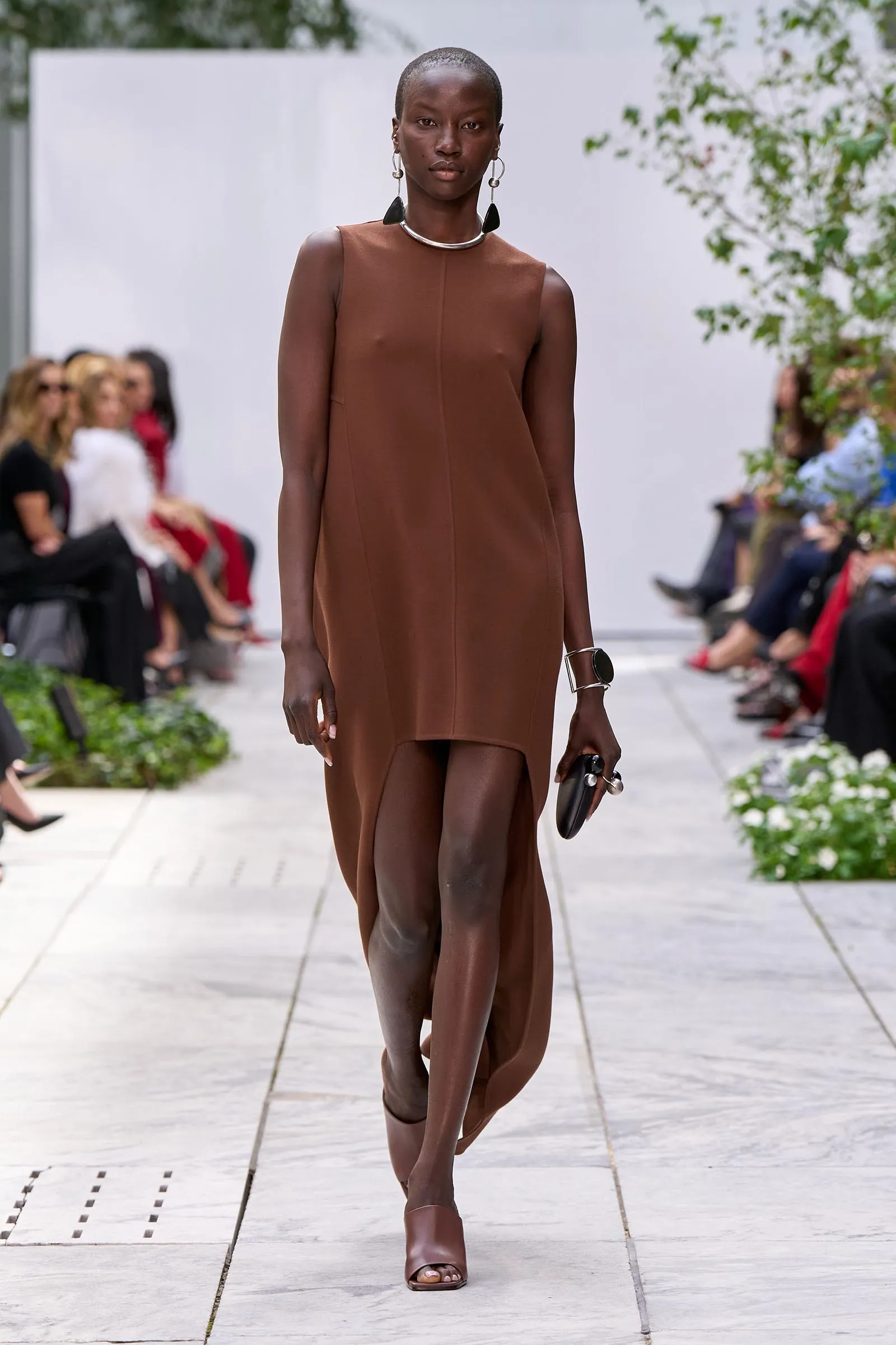 Model walking the runway in a brown sleeveless dress with bold earrings at an outdoor fashion show.