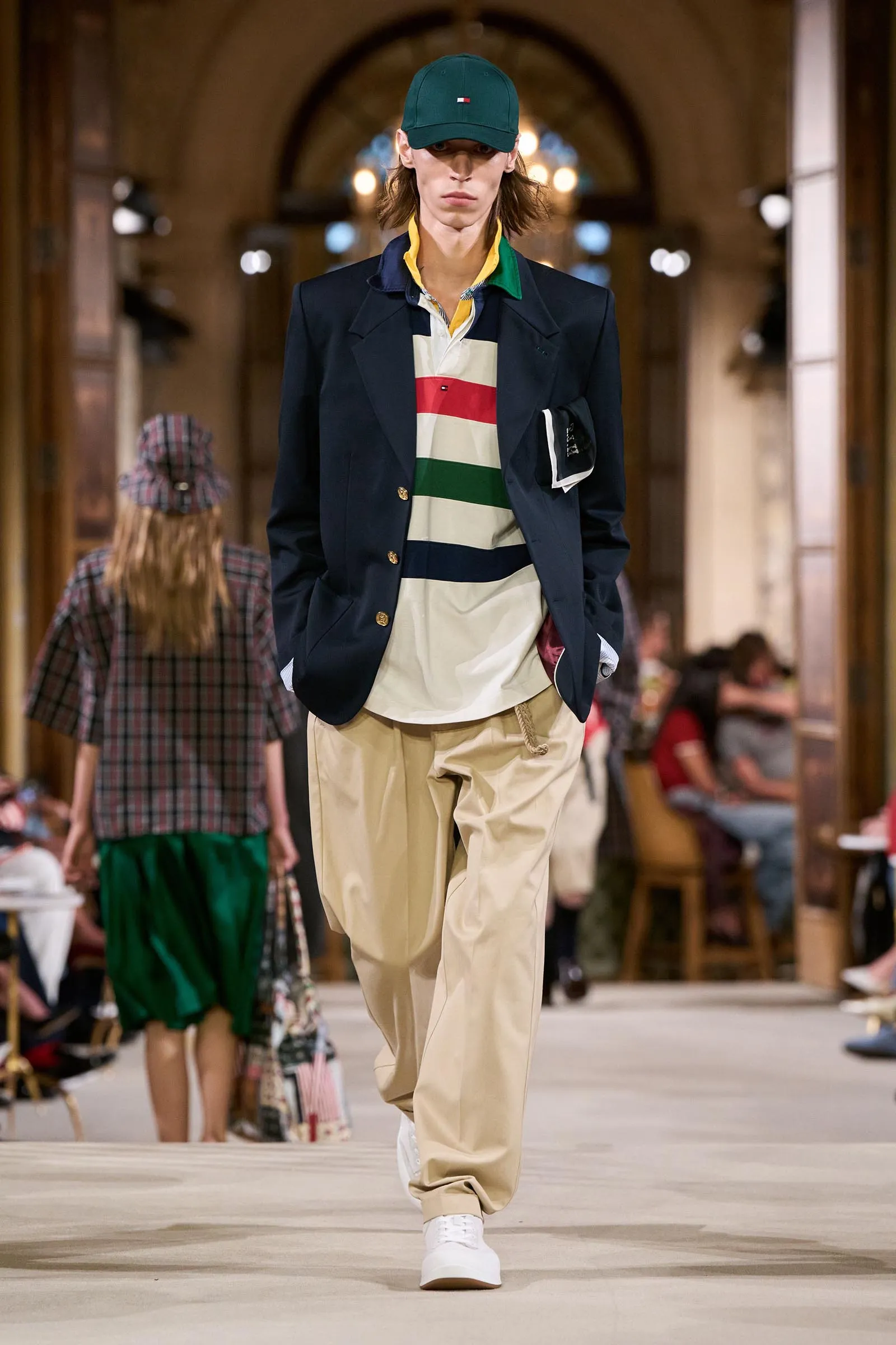 Model walking down a runway in a navy blazer over a striped polo, beige cargo pants, white sneakers and a green cap; audience blurred in the background.