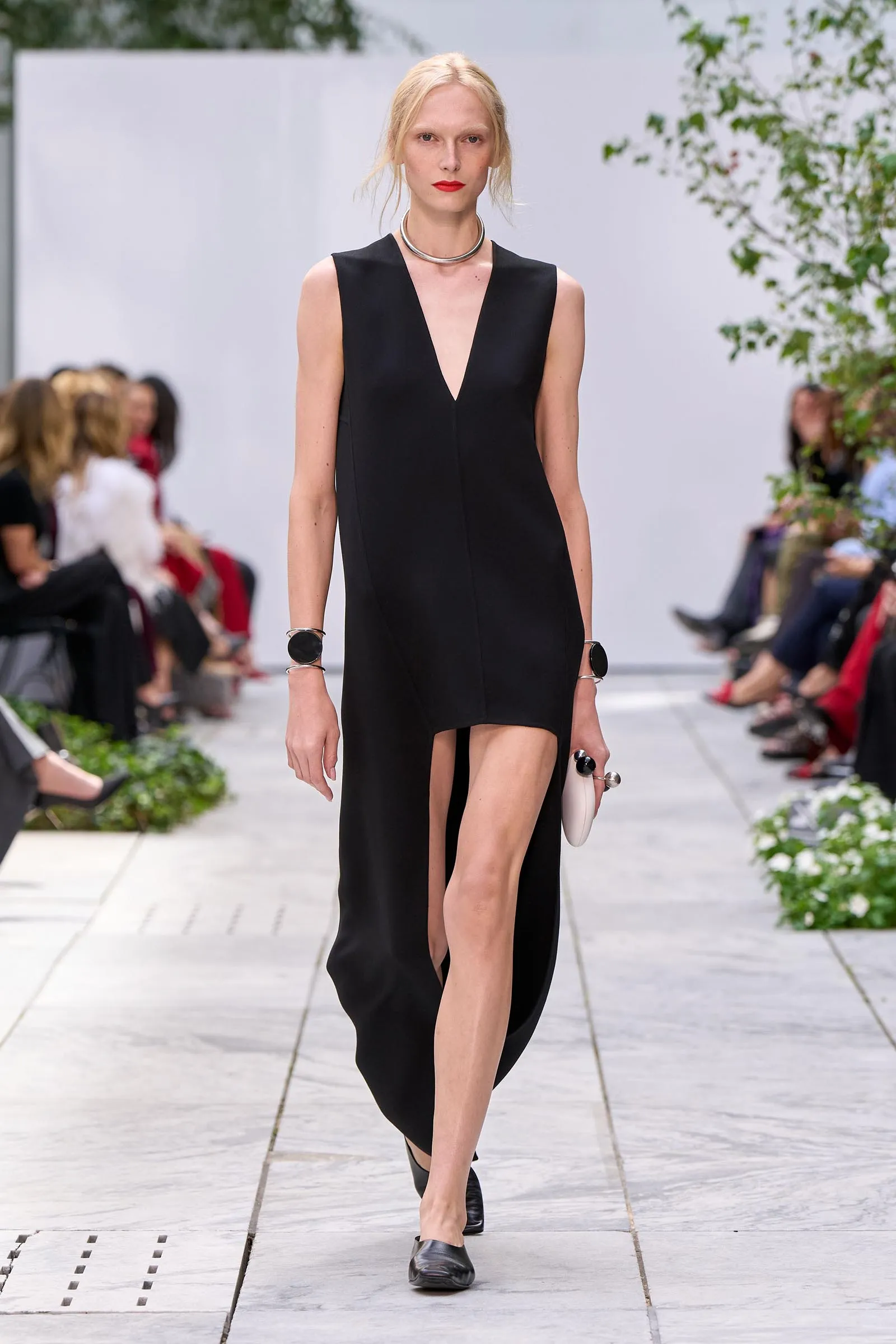 Model walking a runway in a black sleeveless V-neck dress with a high front slit, outdoors with greenery and seated audience in background (fashion show).