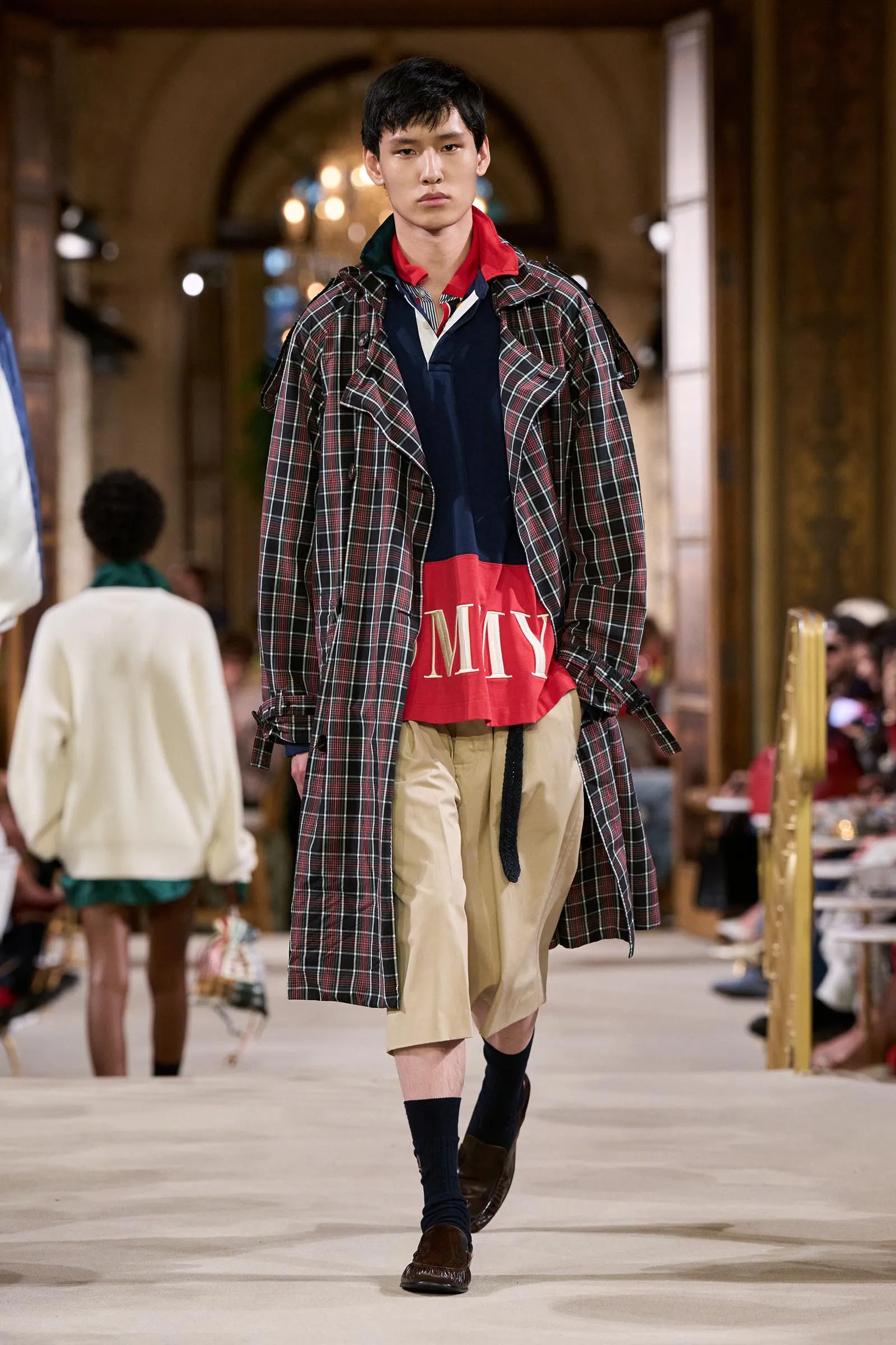 Male model walks the runway in a long plaid trench over a red- and navy-color block top, beige shorts, and dark socks in an ornate indoor setting.