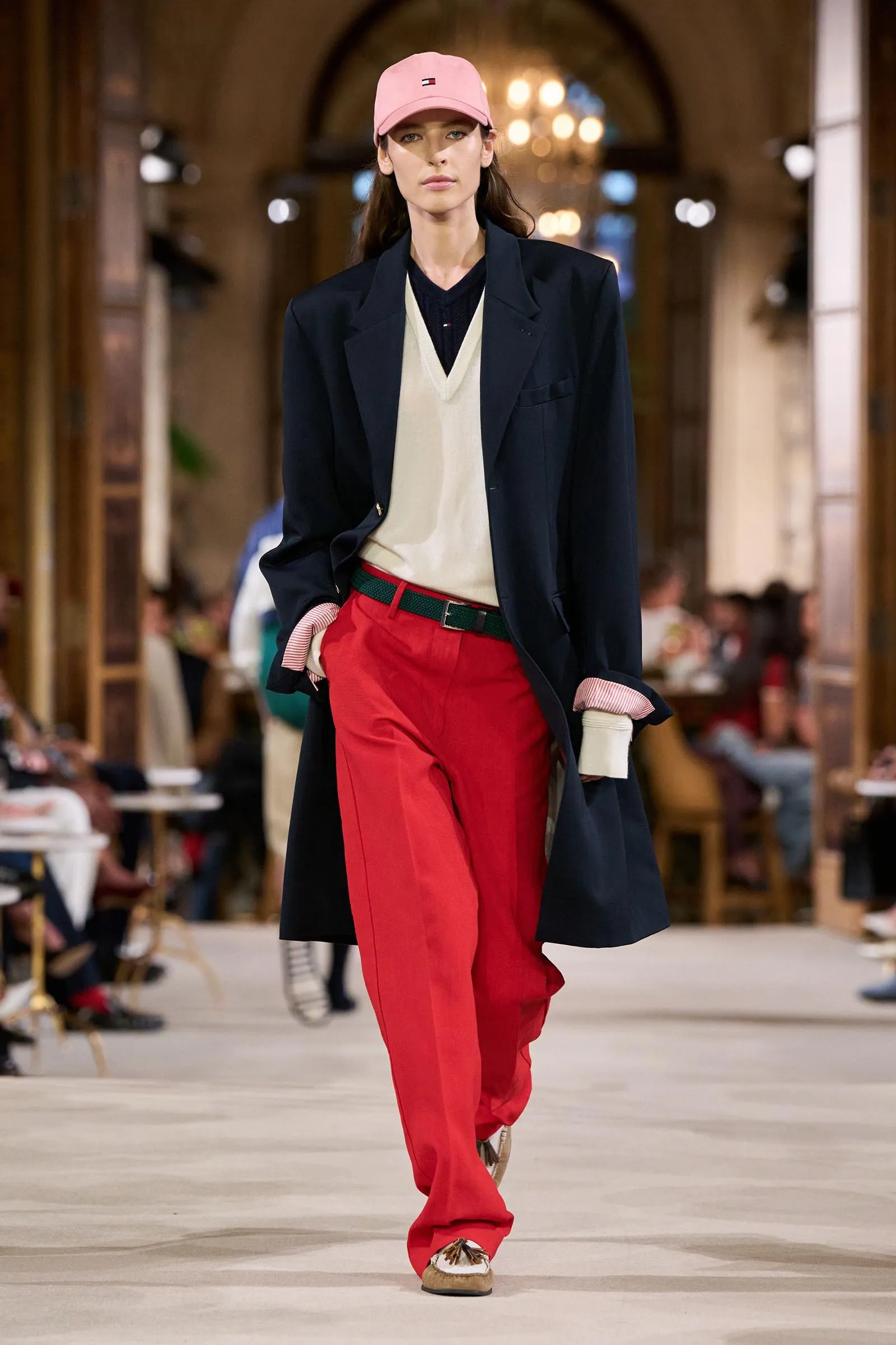 Model on a runway wearing a navy coat, cream sweater, red pants, and a pink cap; blurred audience in the background.