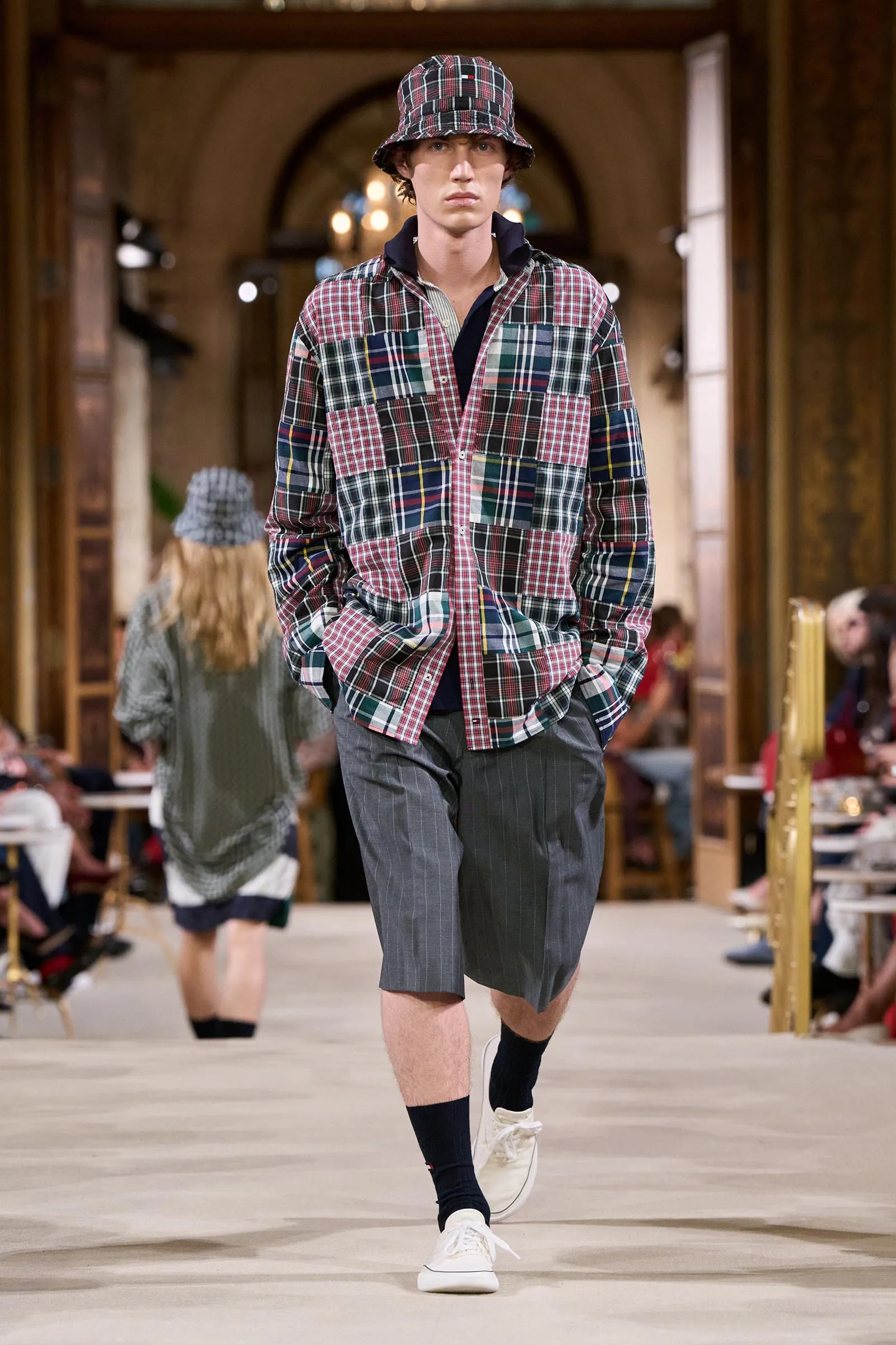 Male model in a multicolored plaid shirt and bucket hat walks a runway in a grand indoor venue with seated spectators in the background.