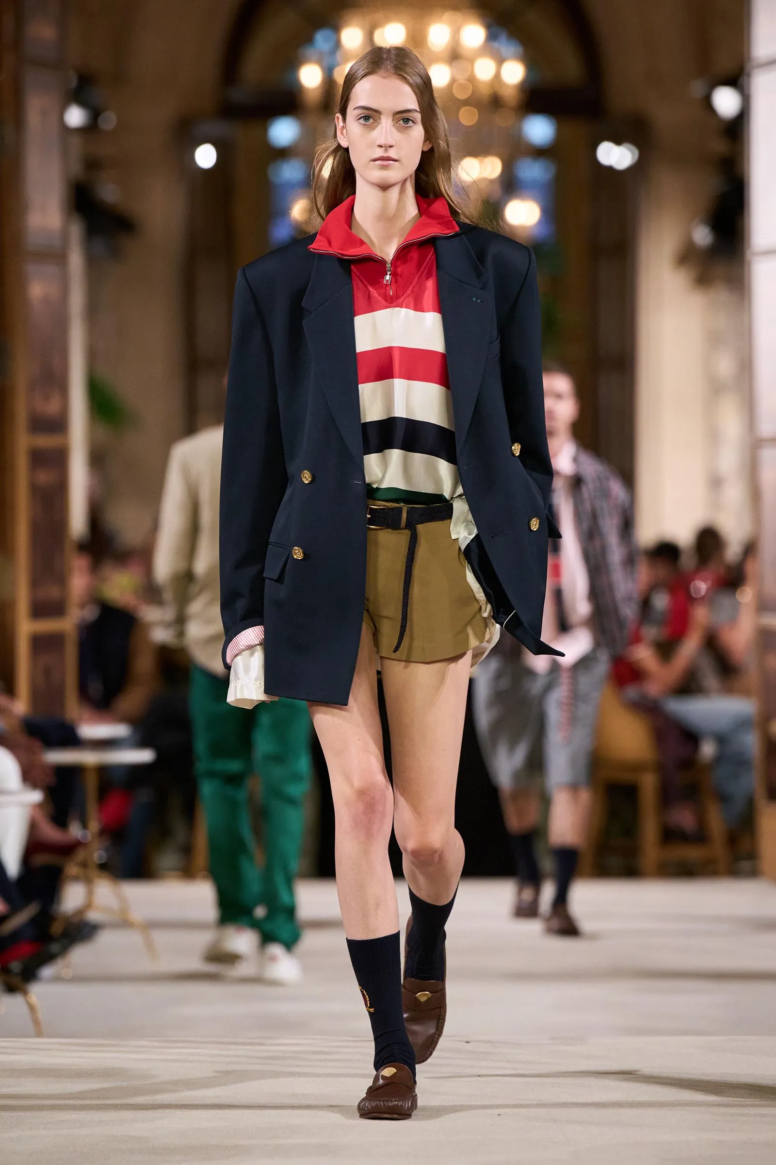 Female model walking a fashion runway in a navy blazer over a red, cream, and black striped top with tan shorts and brown loafers; blurred audience in background.