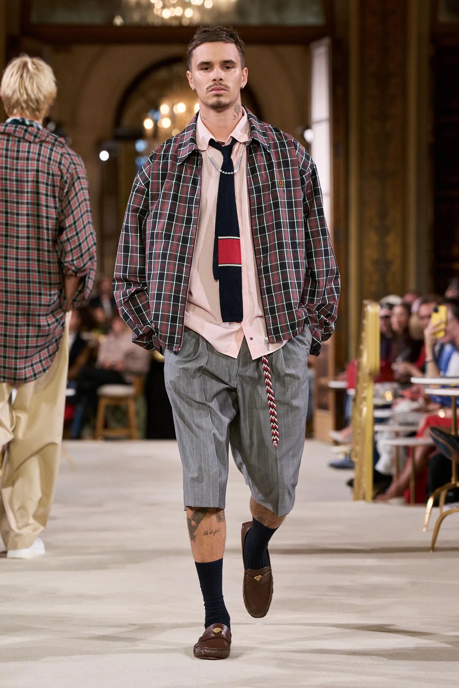 Male model walks the runway in a red‑green plaid oversized jacket over a pale pink shirt and navy tie, gray pinstripe shorts, and loafers in a fashion show venue.