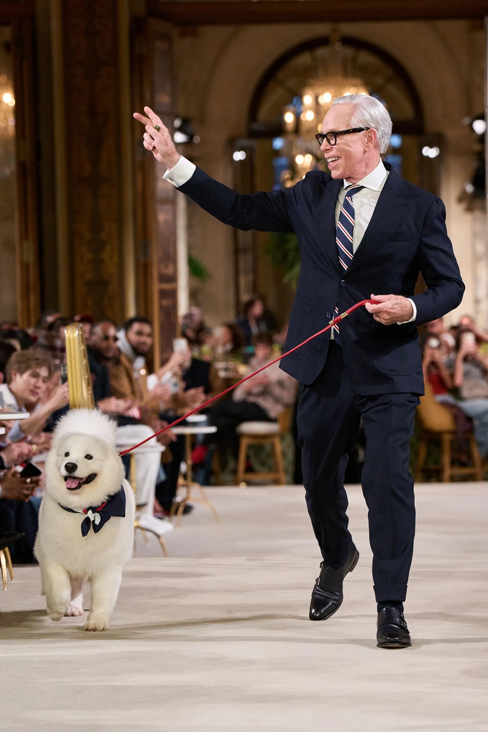 Elegant older man in a navy suit and striped tie leads a white fluffy dog on a red leash along a runway as spectators applaud.