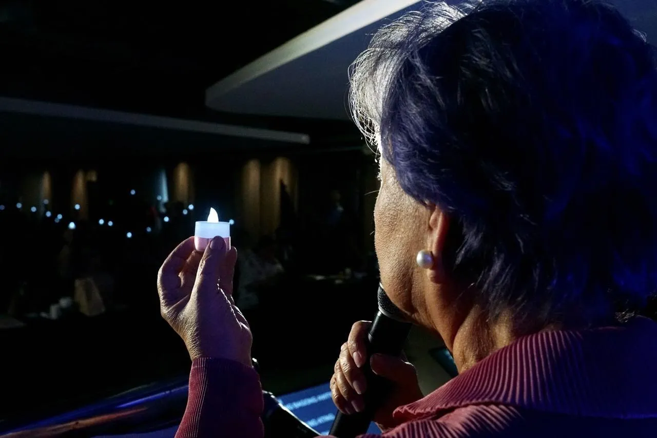 Speaker on a dimly lit stage holds a lit small candle in one hand and speaks into a handheld microphone, with an audience faintly visible in the background.