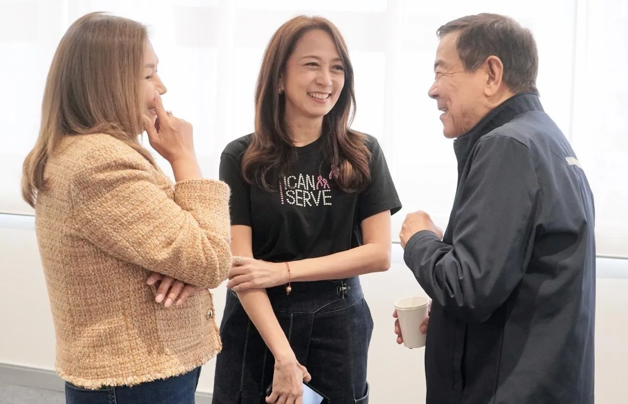 Three colleagues smiling and talking in a bright office, one holding a paper cup.