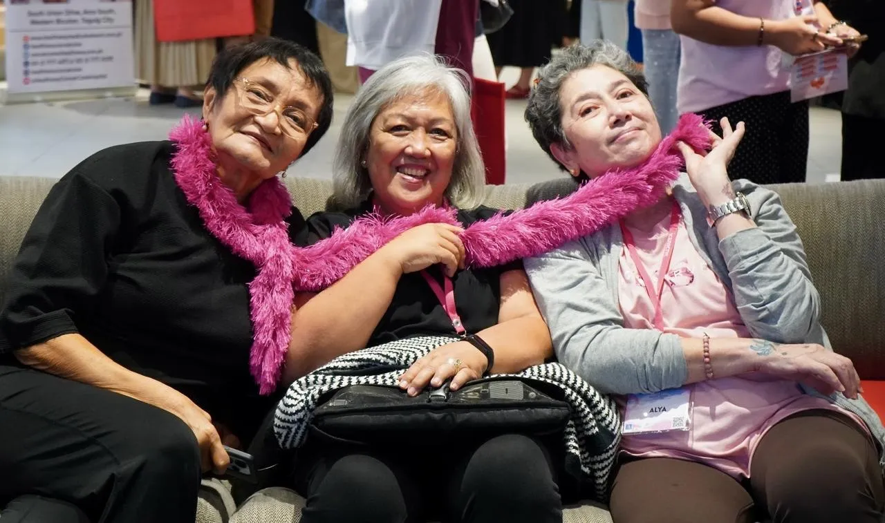 Three women sit close on a sofa, smiling, with a pink feather scarf draped around their shoulders.