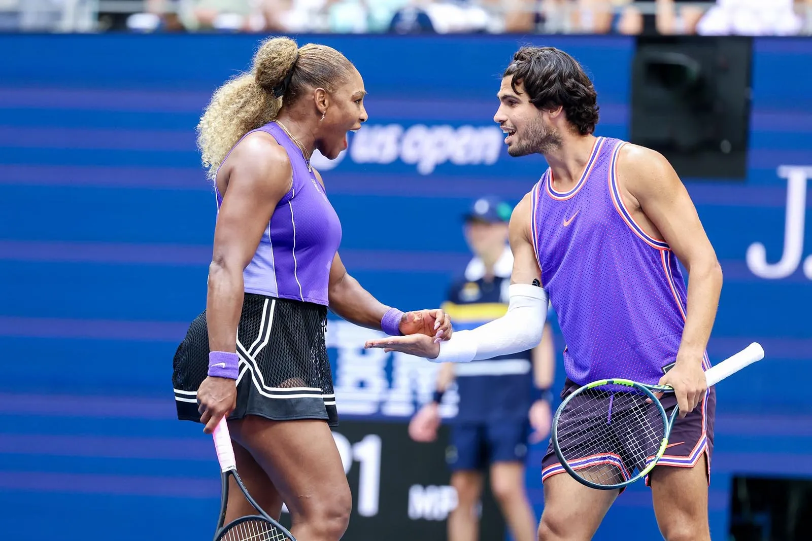 Two tennis players in purple outfits greet each other with a fist bump on court during a match.