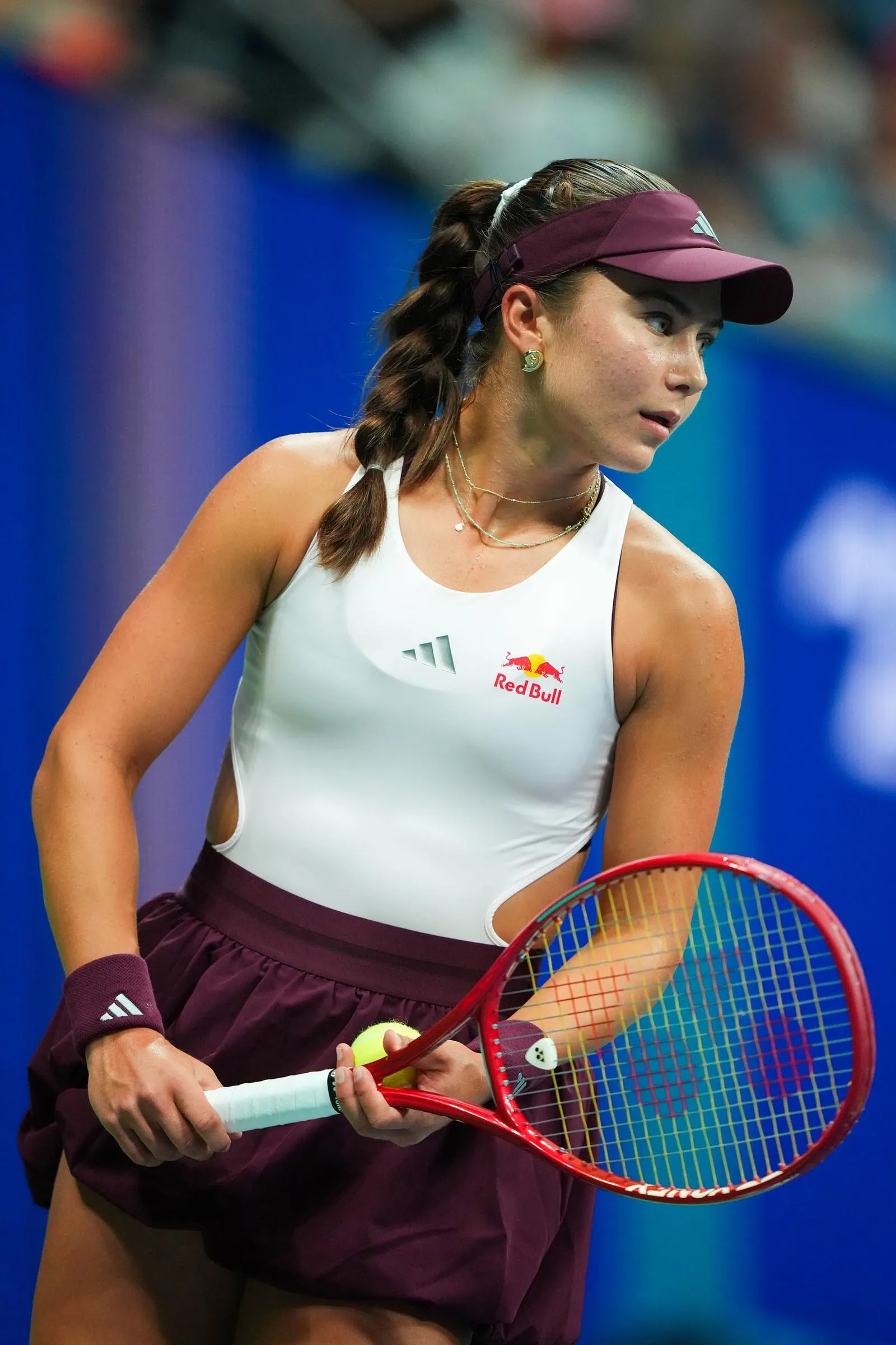 Female tennis player, wearing a burgundy visor and white top with Red Bull logo, holding a red racket and tennis ball.