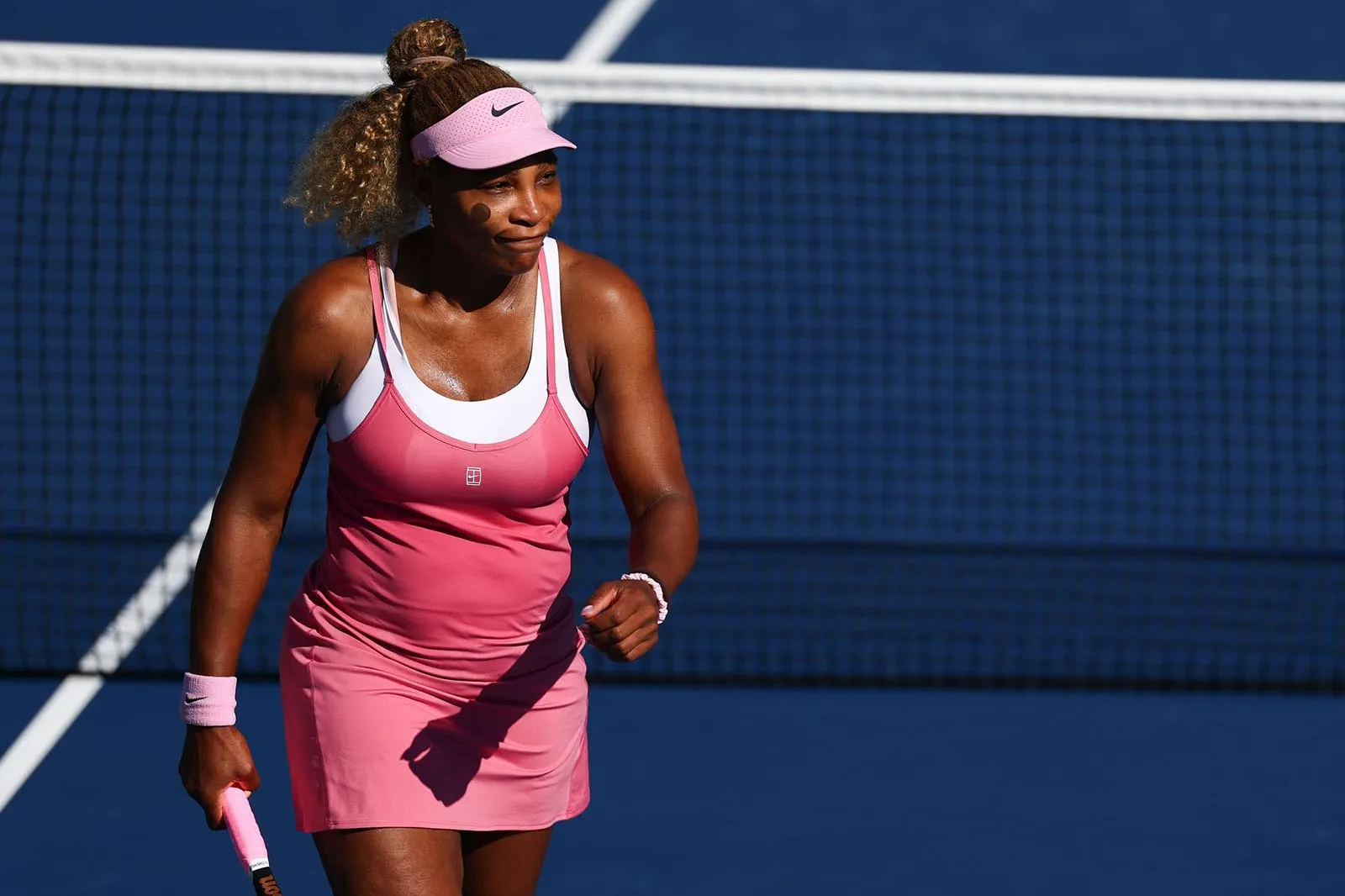 Female tennis player in a pink dress and visor on a blue court, holding a racket and ready to play.