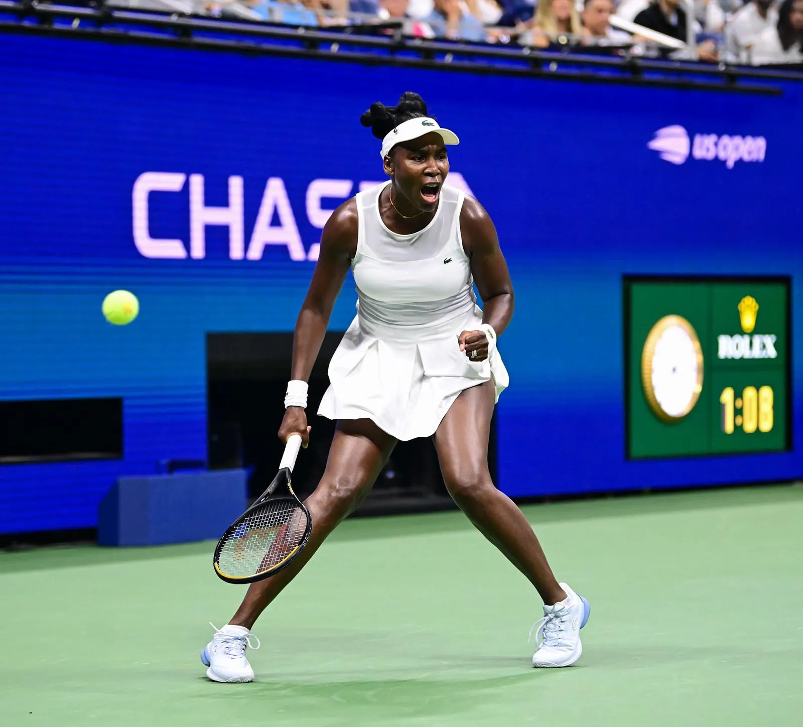 Female tennis player in a white outfit lunges to hit a forehand as a tennis ball approaches on a green hard court, with a blue stadium backdrop and spectators in the background.