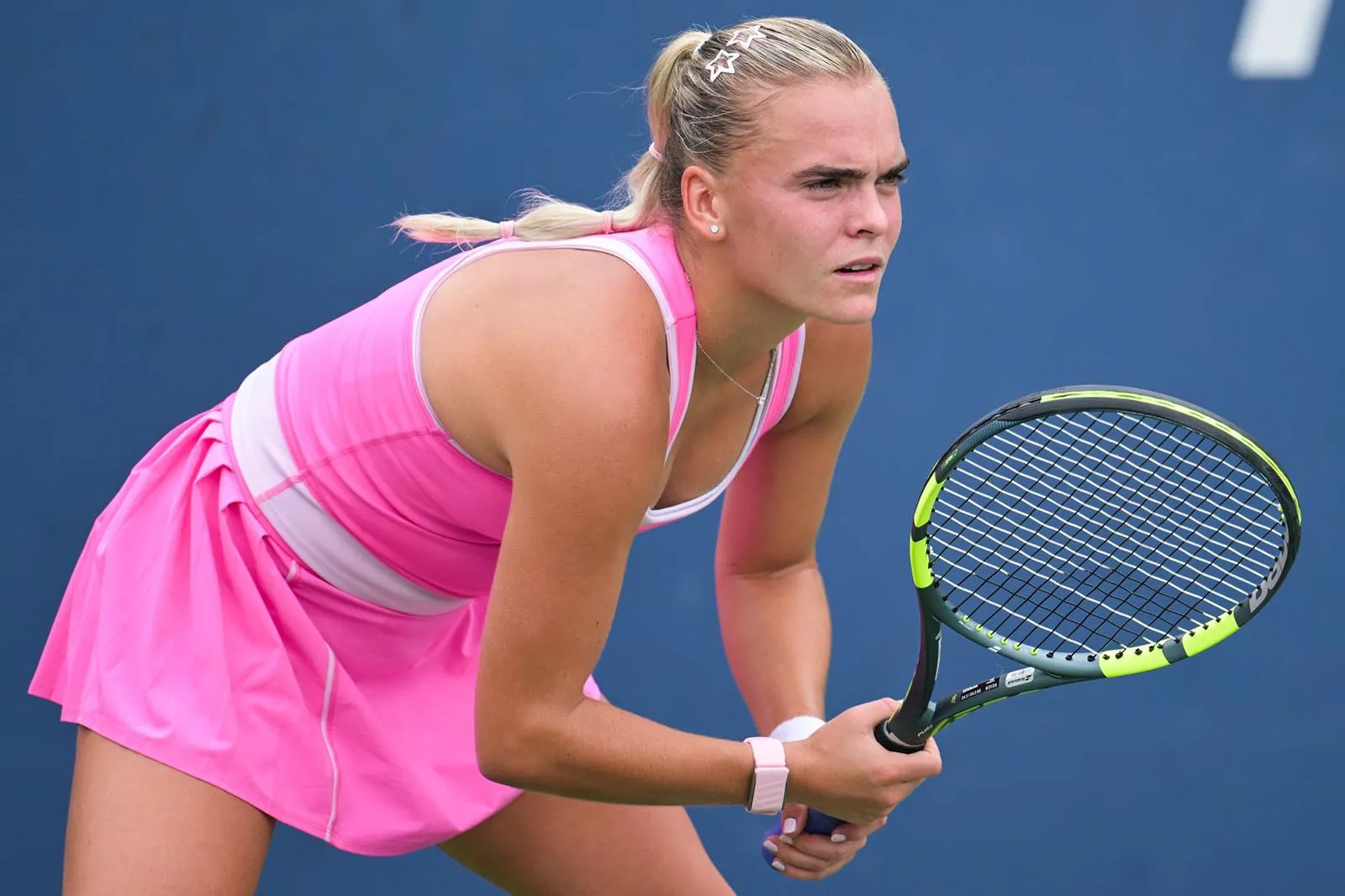 Female tennis player in a pink outfit crouches in a ready stance, holding a racket on a blue court.