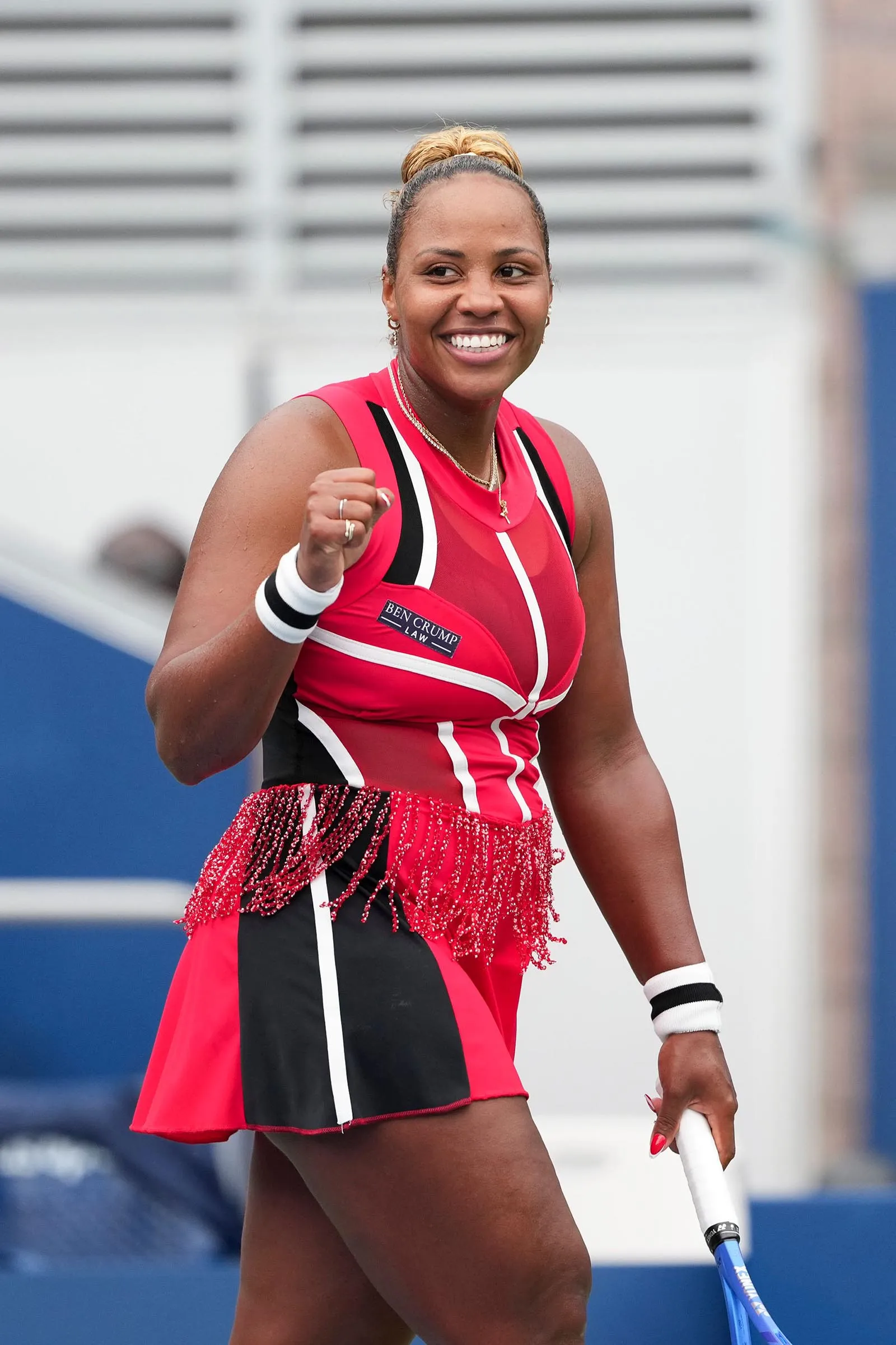 Smiling female tennis player in a red and black outfit raises her fist, holding a blue racket on the court.